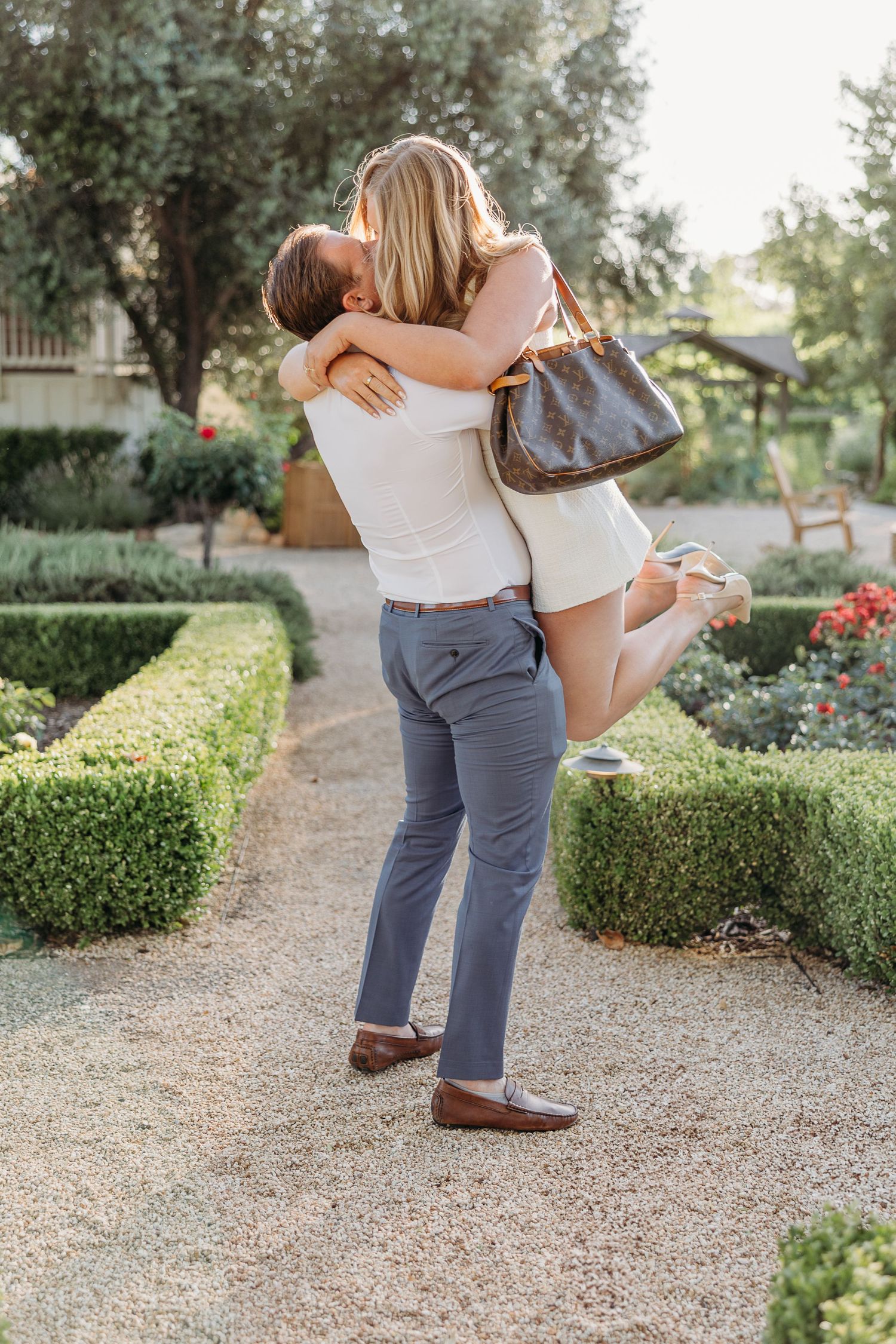A joyful couple embraces in a garden path lined with manicured hedges on a sunny afternoon.