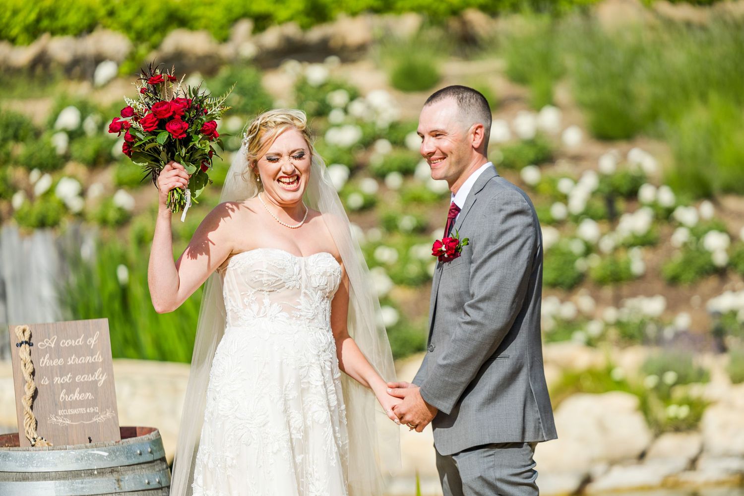 Joyful wedding moment with red bouquet against garden backdrop.