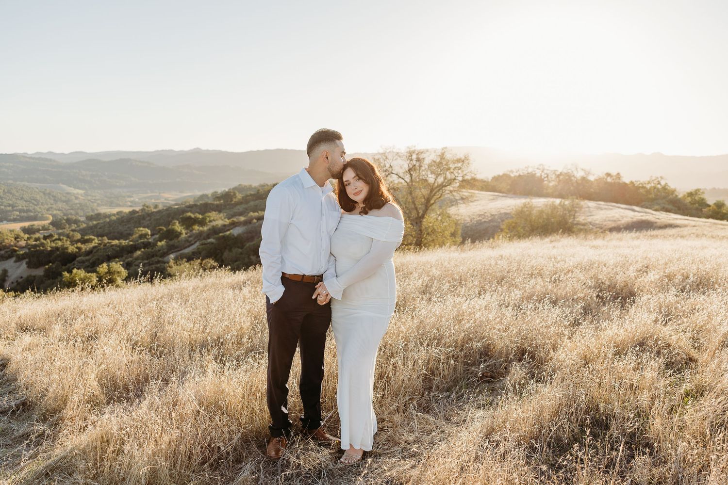 A couple stands together in golden sunlit fields during sunset at a mountain overlook.