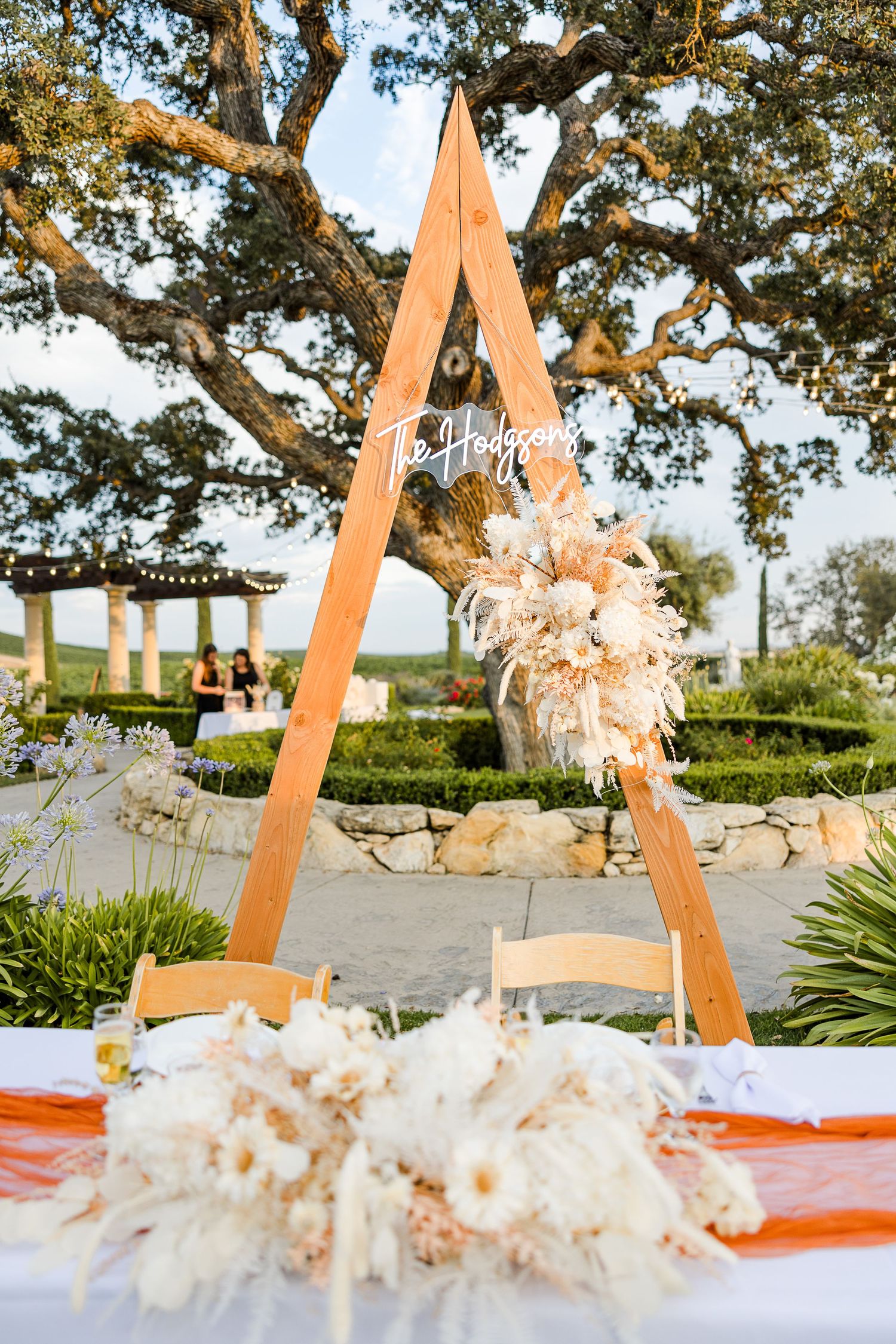 Wooden A-frame wedding arch decorated with dried pampas grass and florals at an outdoor venue.