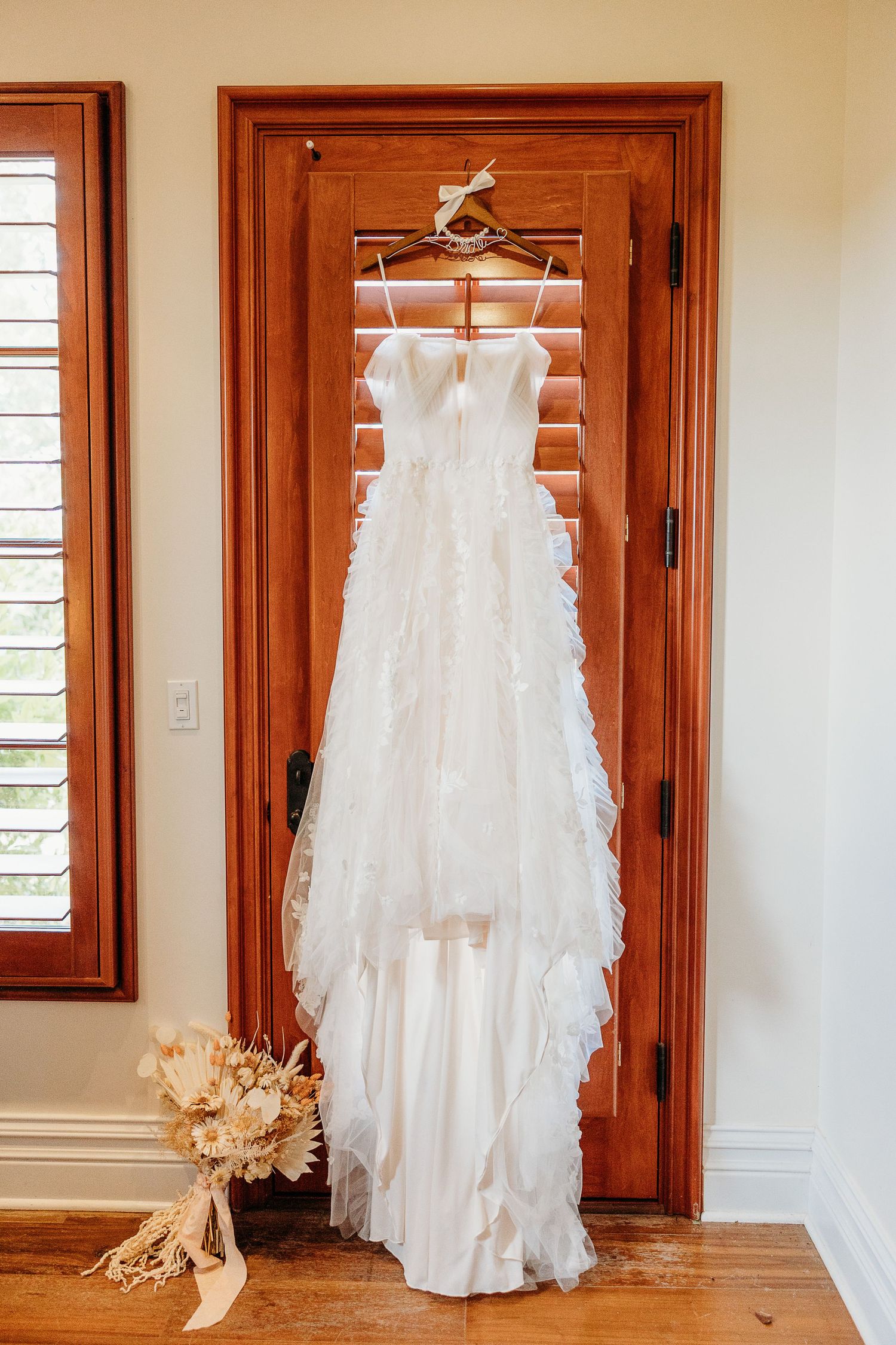 White lace wedding gown hanging on wooden door with natural light streaming through windows.