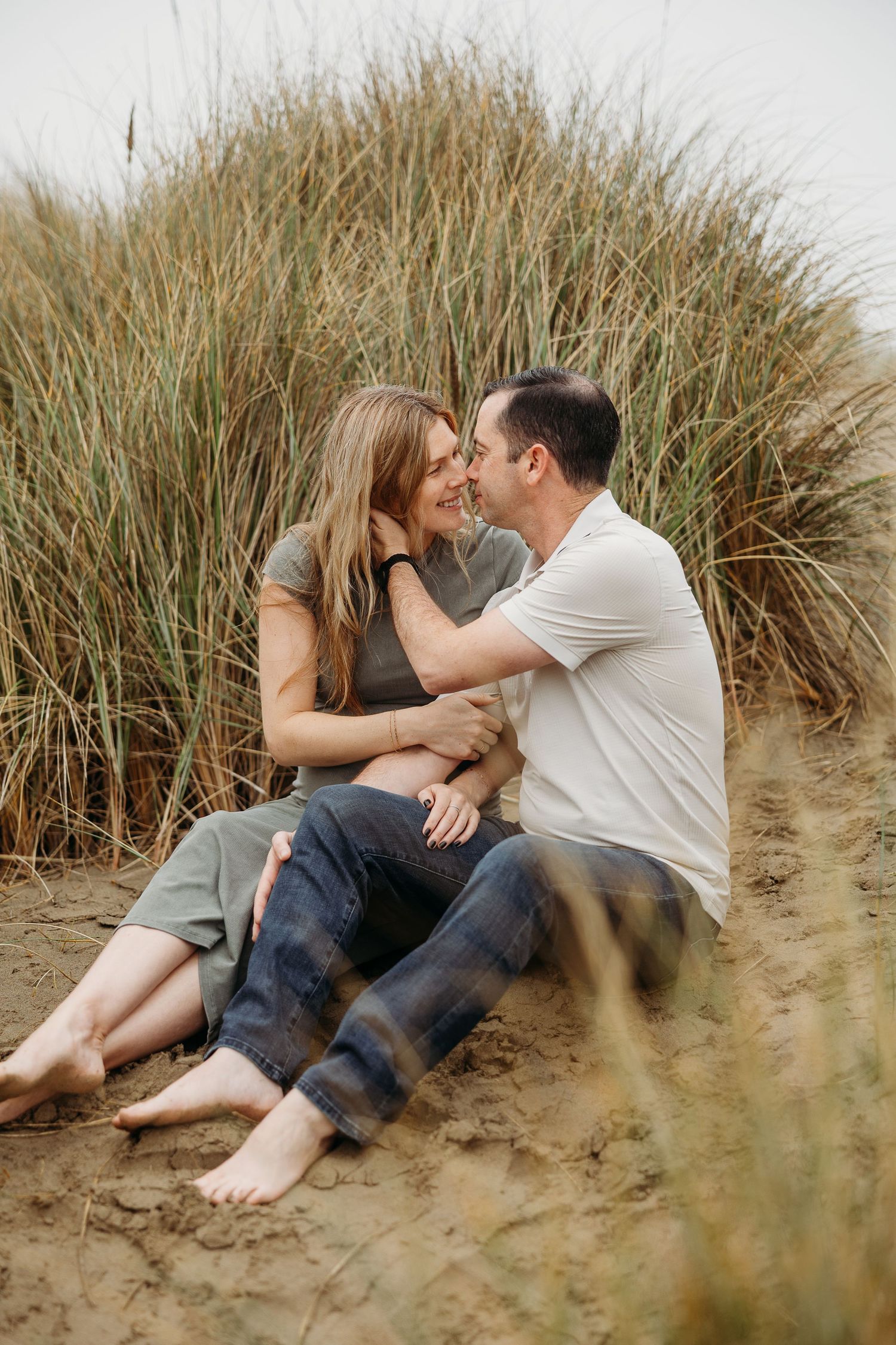 Natural maternity portrait of couple sitting barefoot in sand surrounded by beach grass.