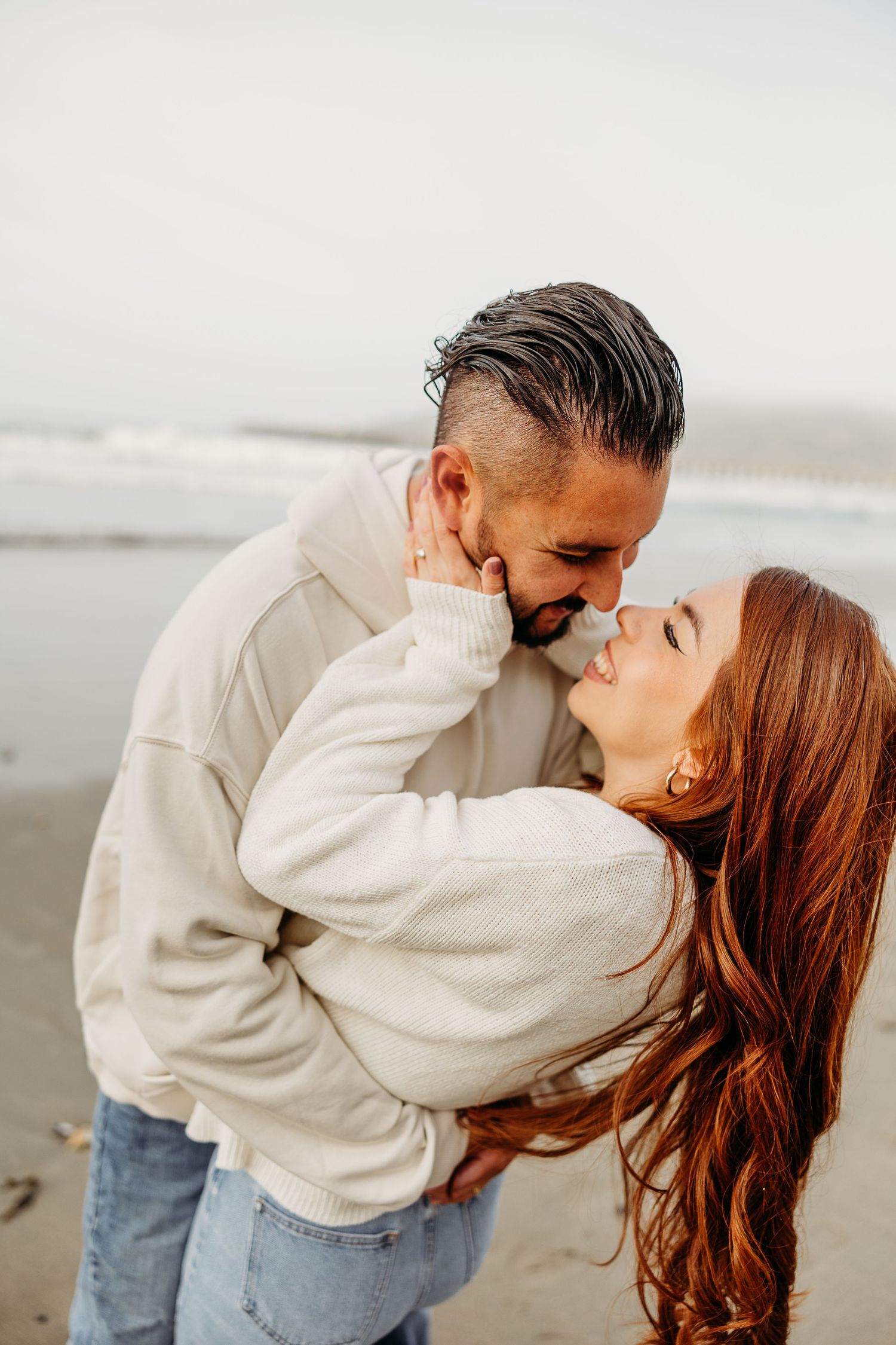 Couple in cozy cream sweaters embracing and kissing on a cloudy beach.