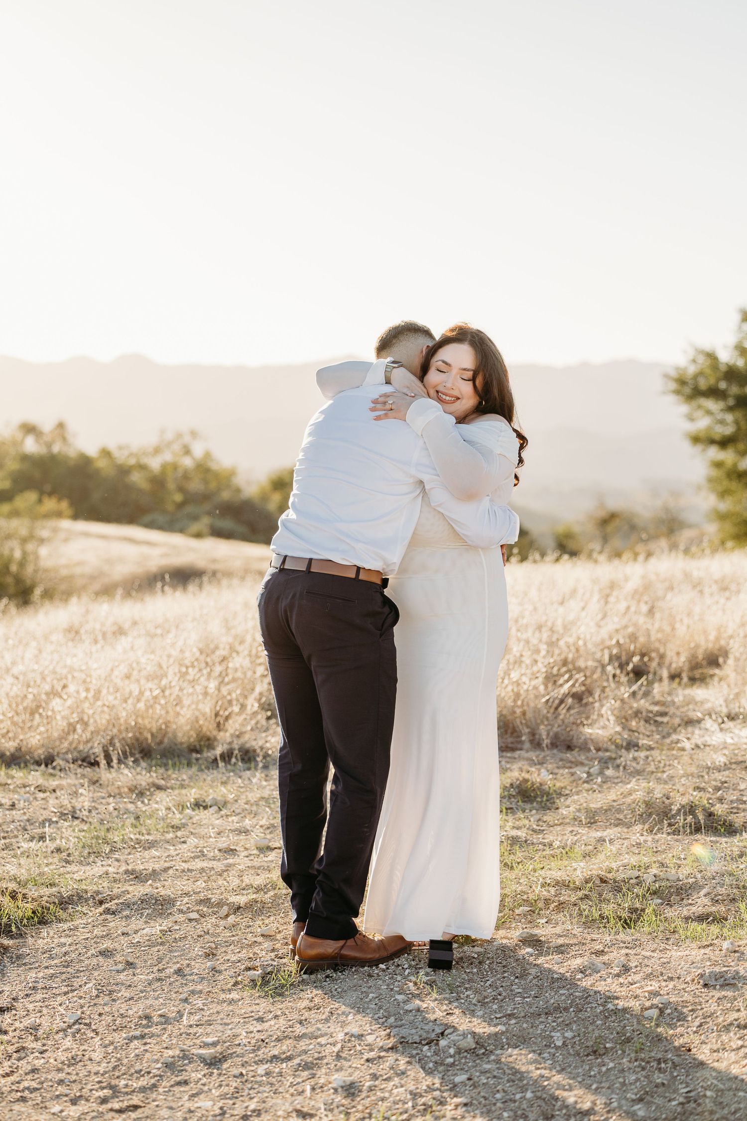 A couple embraces on a sunlit hilltop during what appears to be a proposal moment.