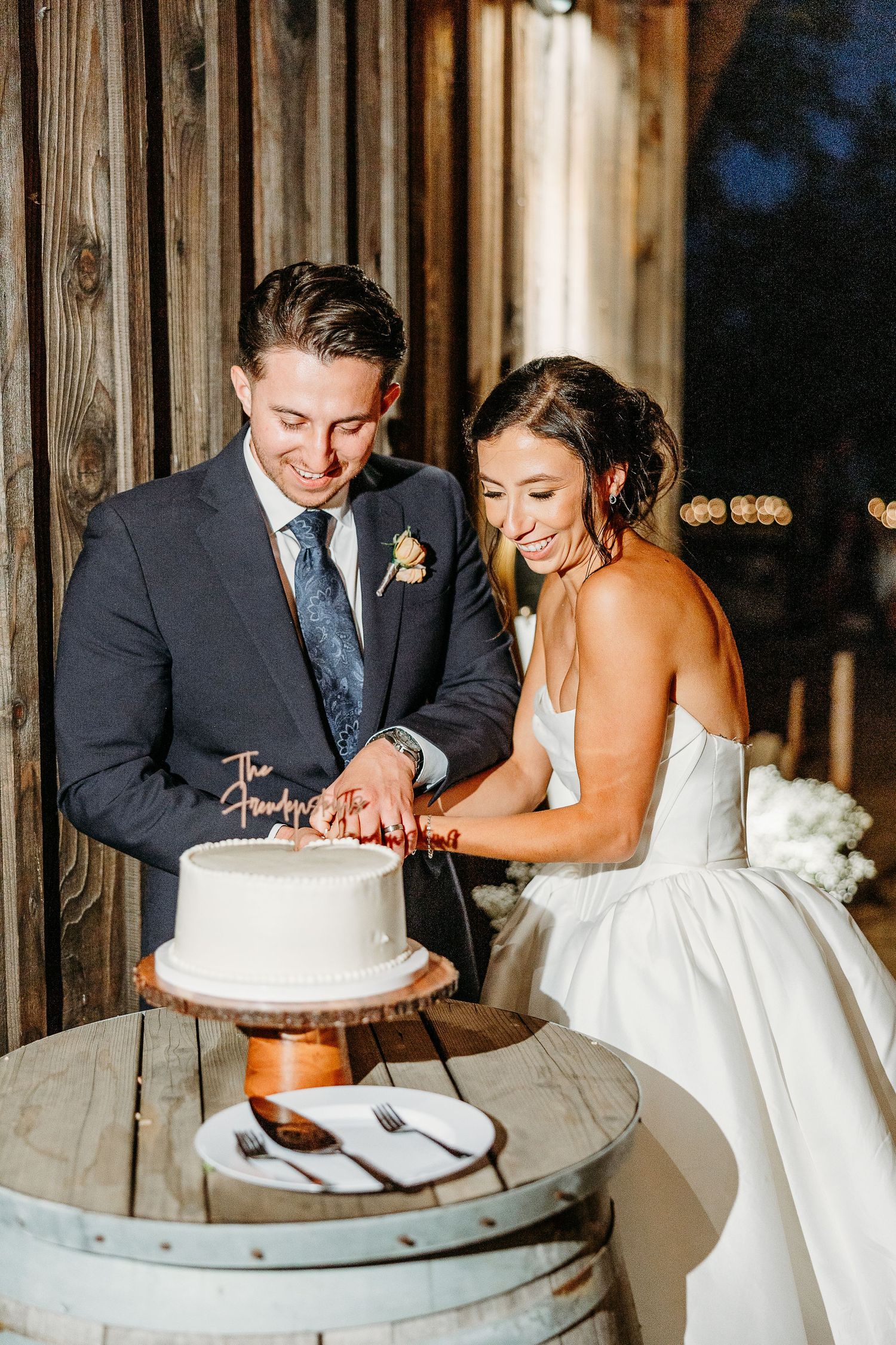 Newlyweds cutting their white wedding cake together at rustic wooden reception venue.