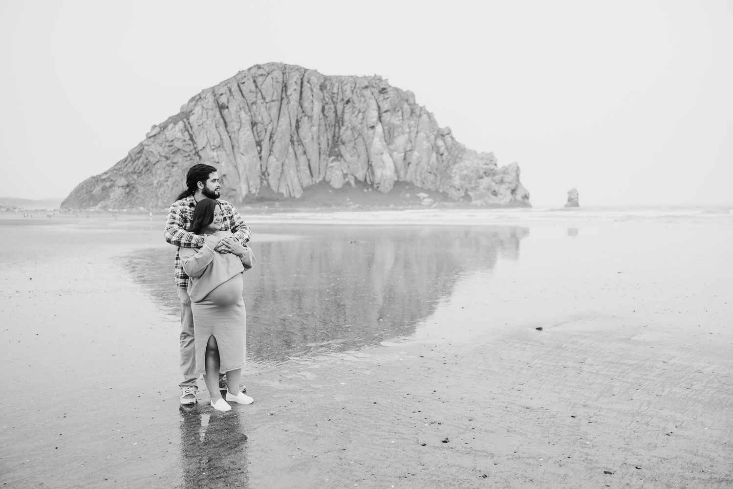 Figure stands on misty beach with iconic Morro Rock formation in background at Morro Bay, California.