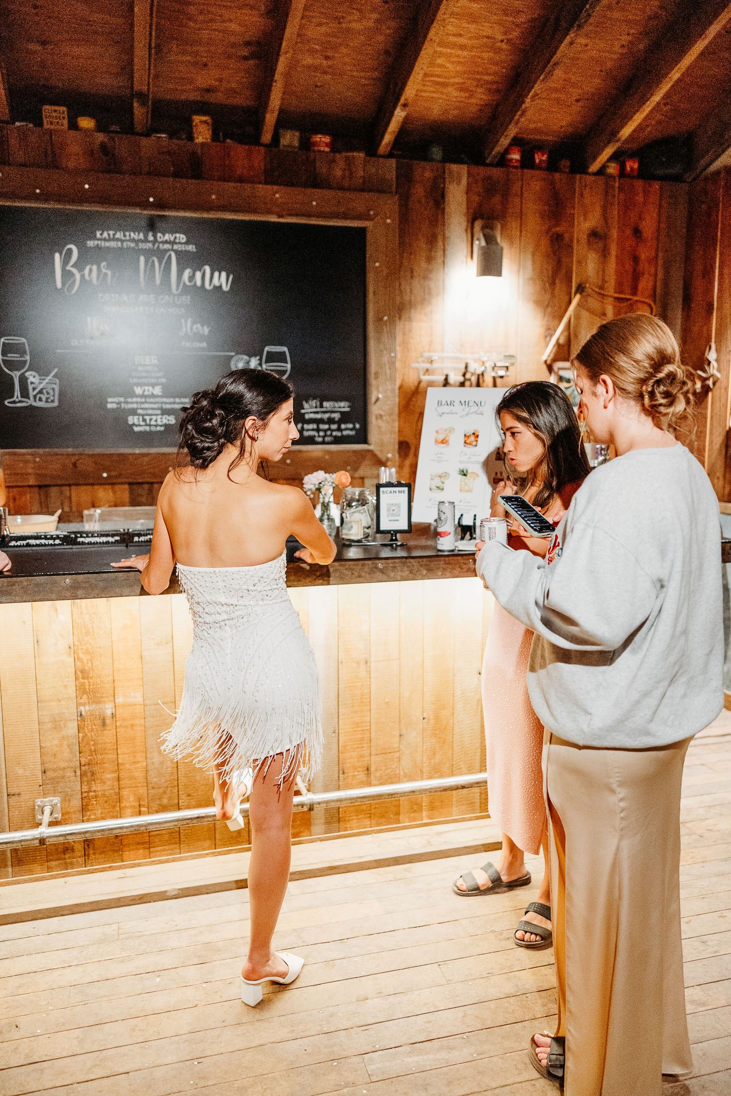 Wedding reception bar setup inside rustic wooden barn with chalkboard menu and ambient lighting.