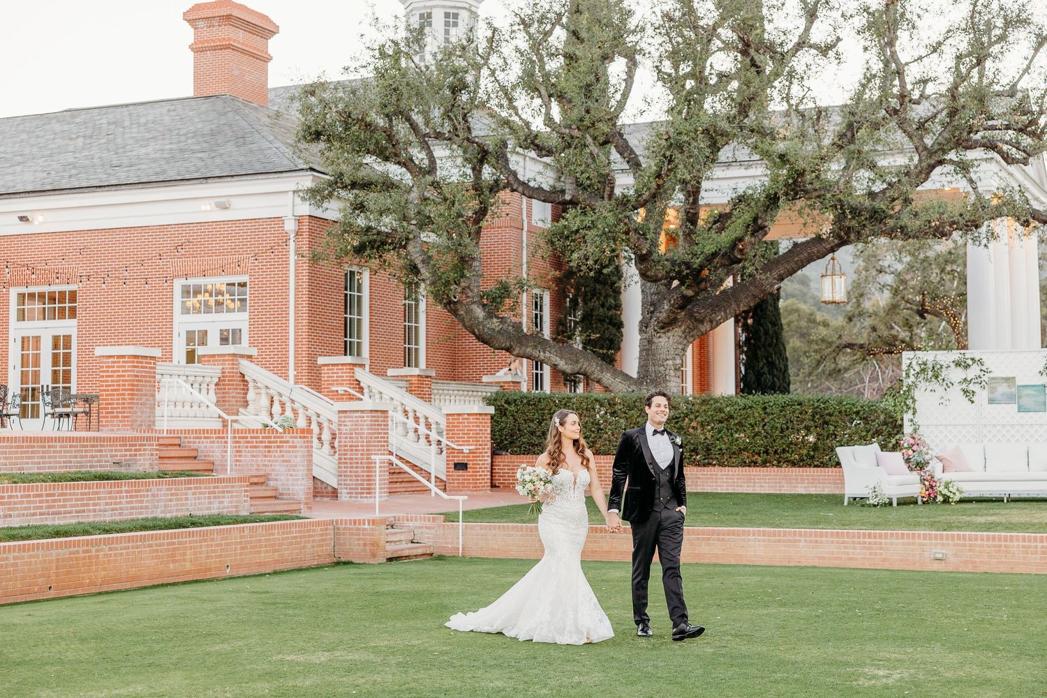 Wedding couple walks across manicured lawn with classic brick building in background.
