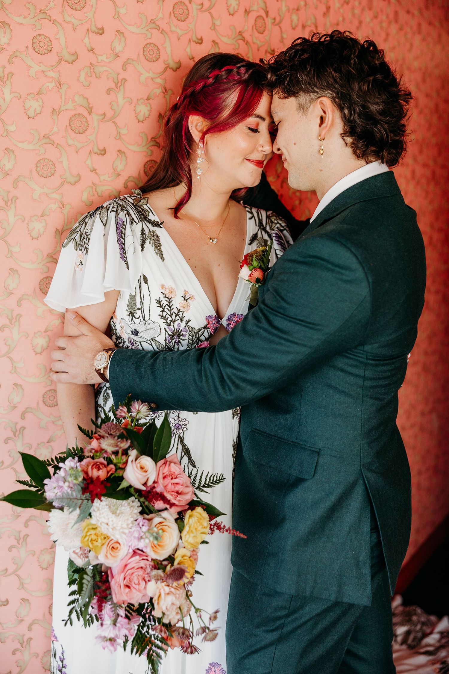 A romantic wedding moment with a floral dress, emerald suit, and vibrant bouquet against a pink patterned wall.