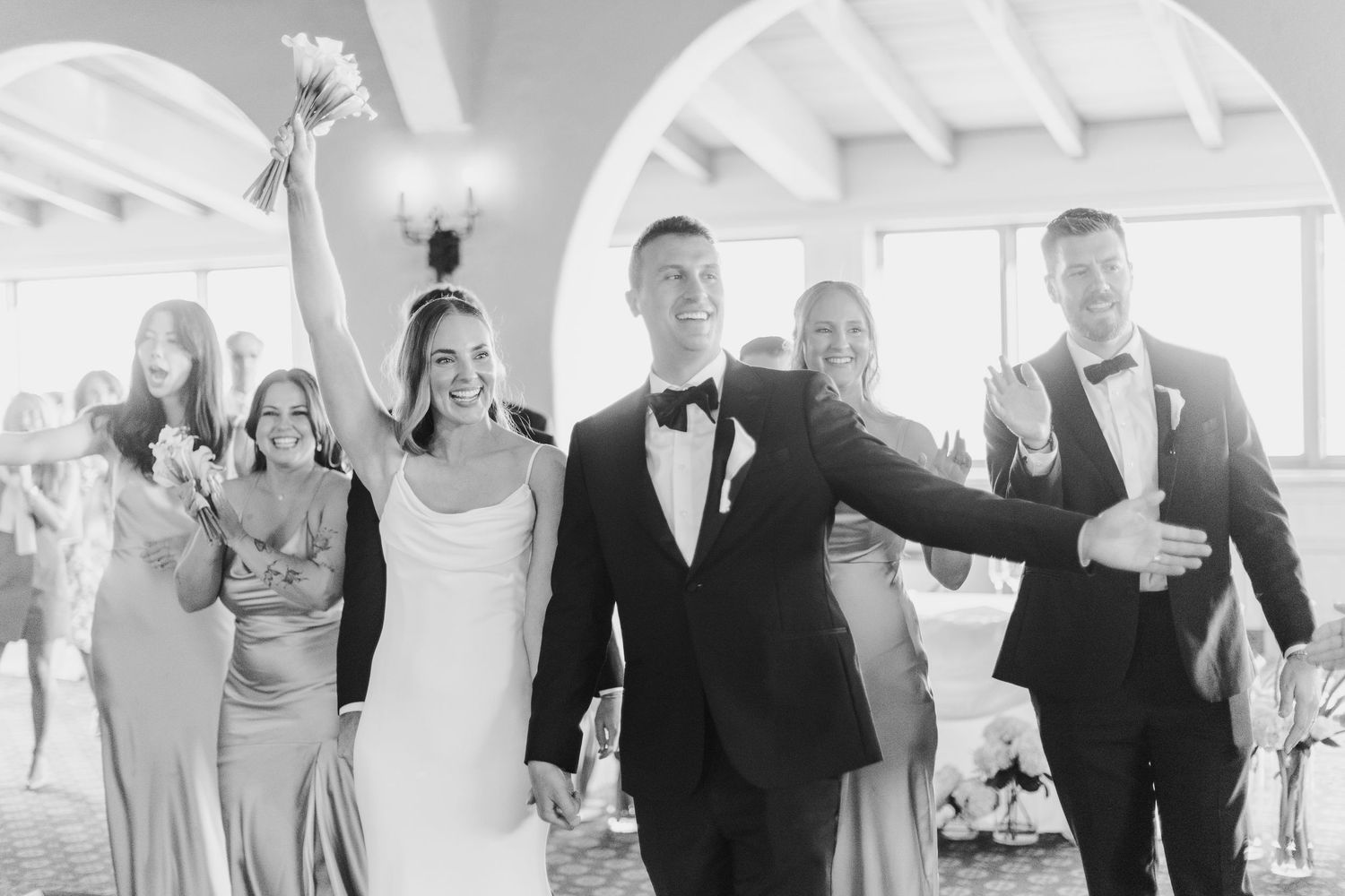 A joyful wedding party celebrates while walking through an elegant arched hallway.