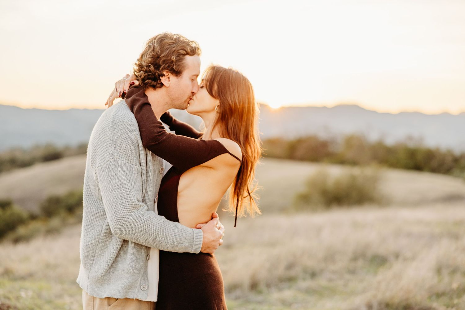 A romantic couple shares a tender moment in a sunlit field with mountains visible in the background.
