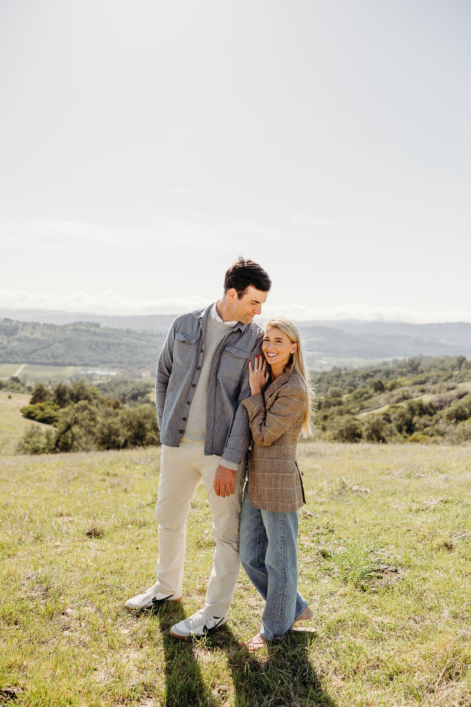 Couple sharing tender moment in sunny meadow with rolling hills behind them.