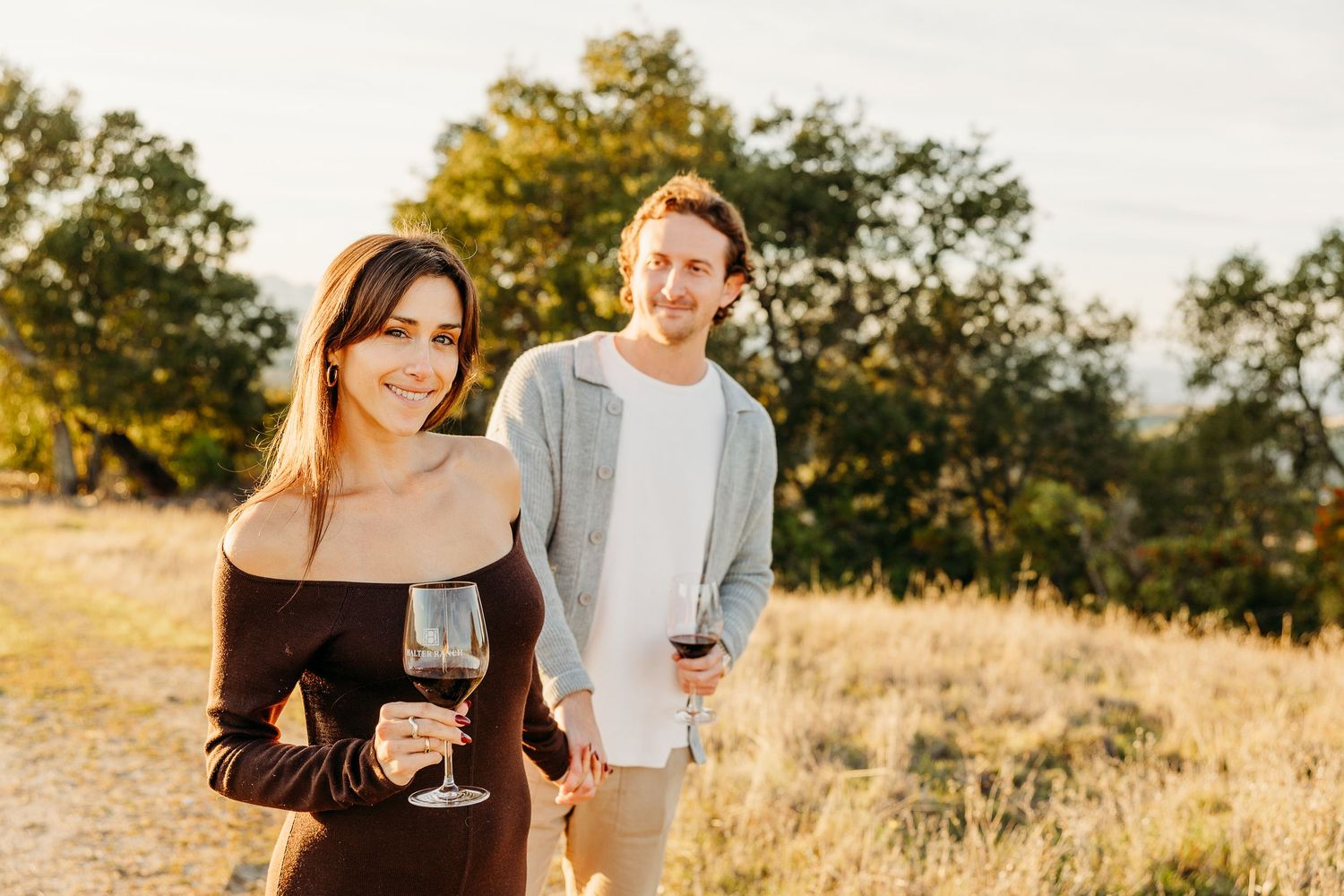 Couple toasting with red wine glasses against golden sunset landscape.