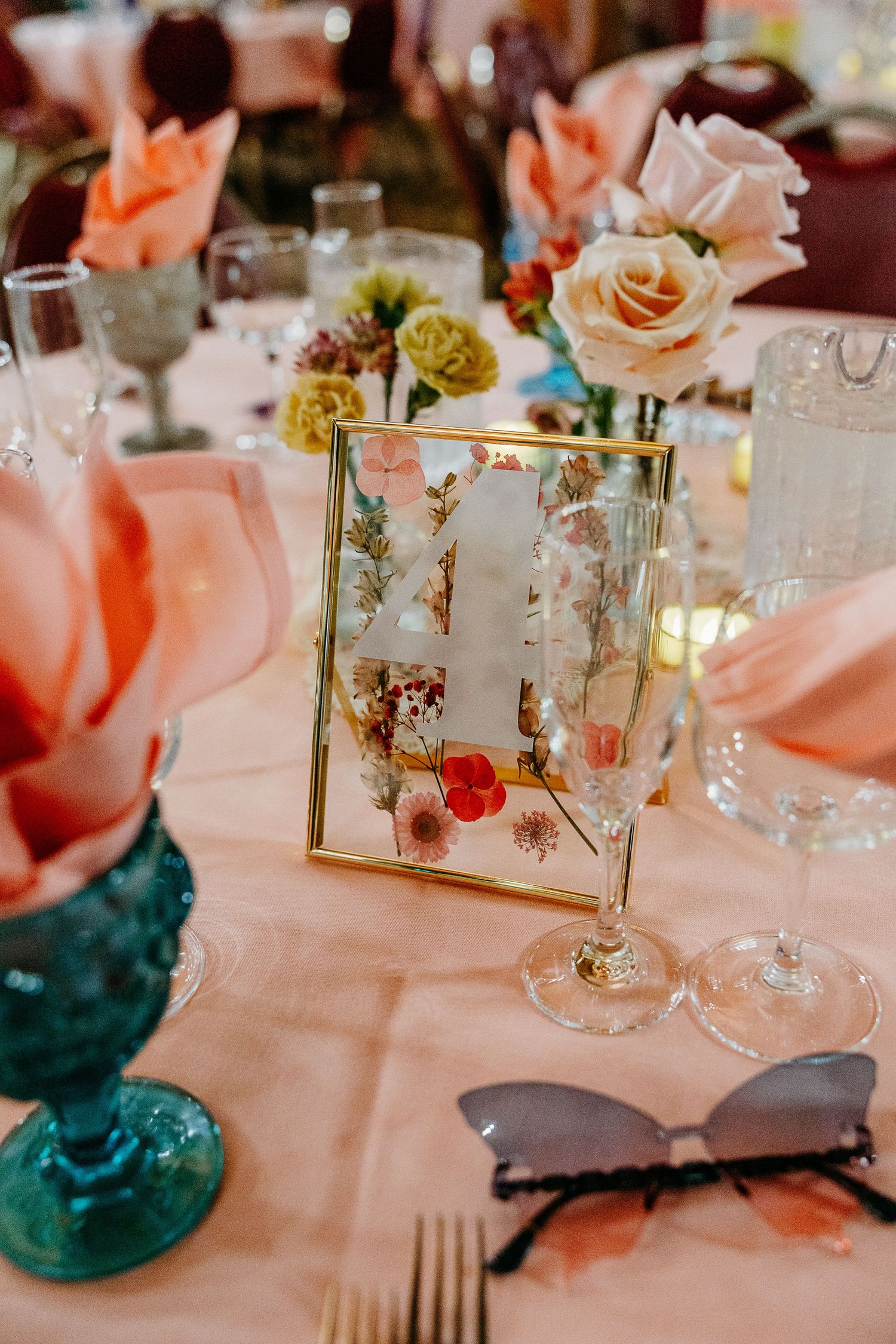 Elegant wedding reception table with pink roses, teal glasses, and gold frame decorations.