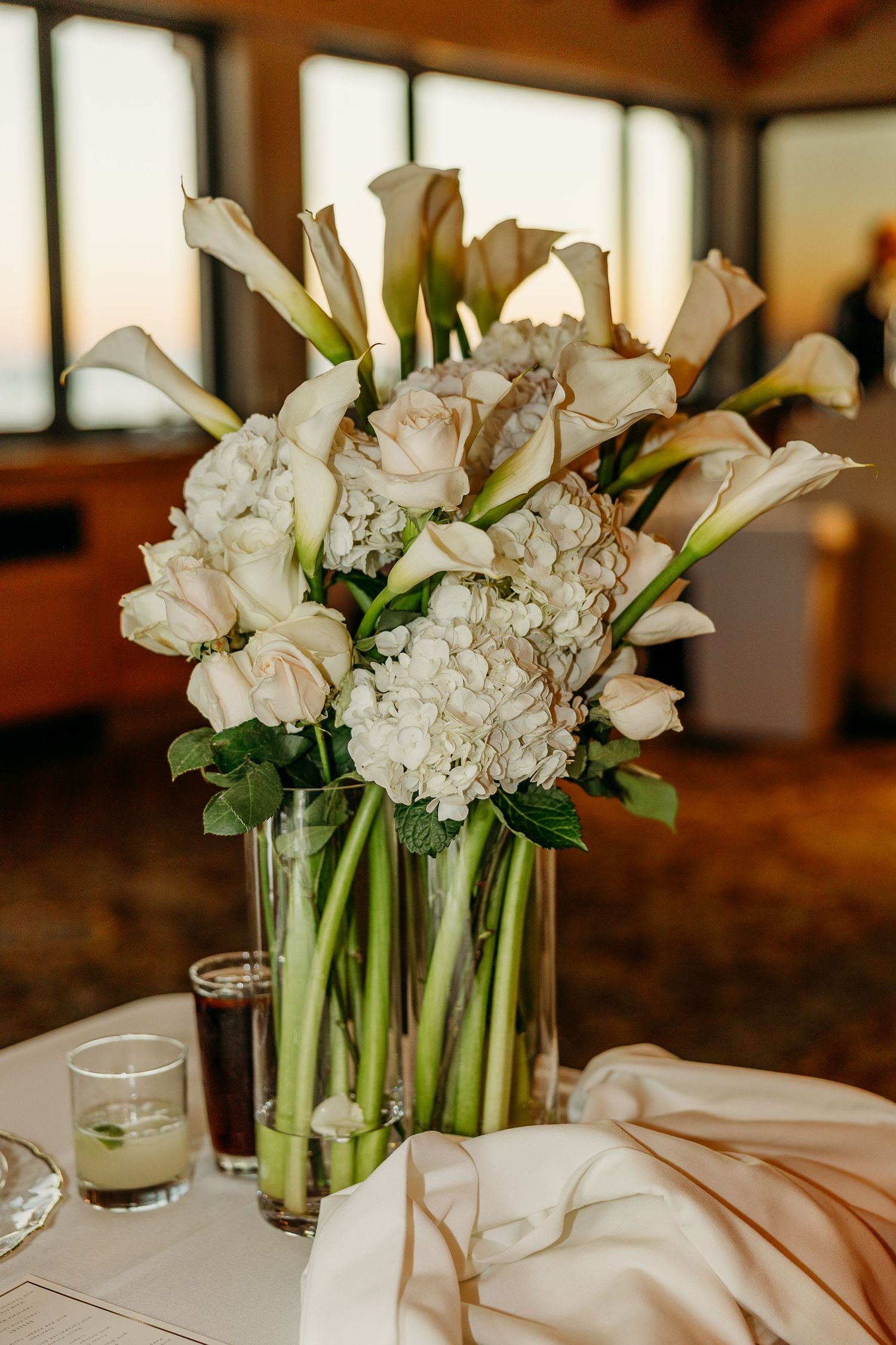Elegant white floral centerpieces with calla lilies and hydrangeas on reception tables.