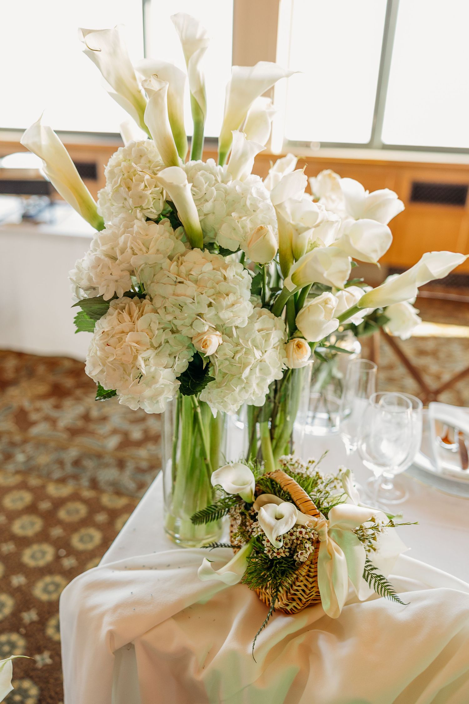 Elegant white floral centerpieces with calla lilies and hydrangeas on tables at a wedding reception.