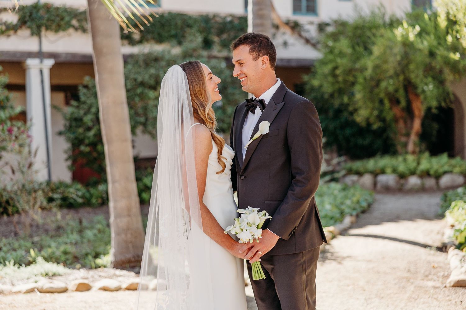 The newlyweds share an intimate moment in a sun-drenched garden with palm trees and Spanish architecture.