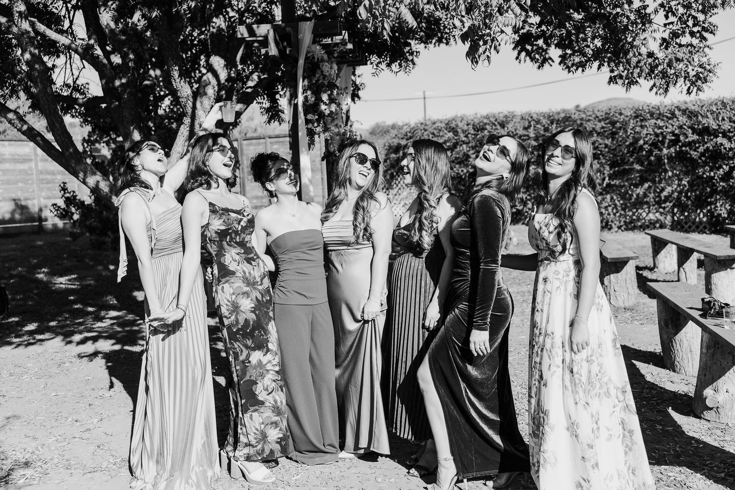 Wedding party members in elegant dresses and suits gather outdoors for group photos at a rustic venue.