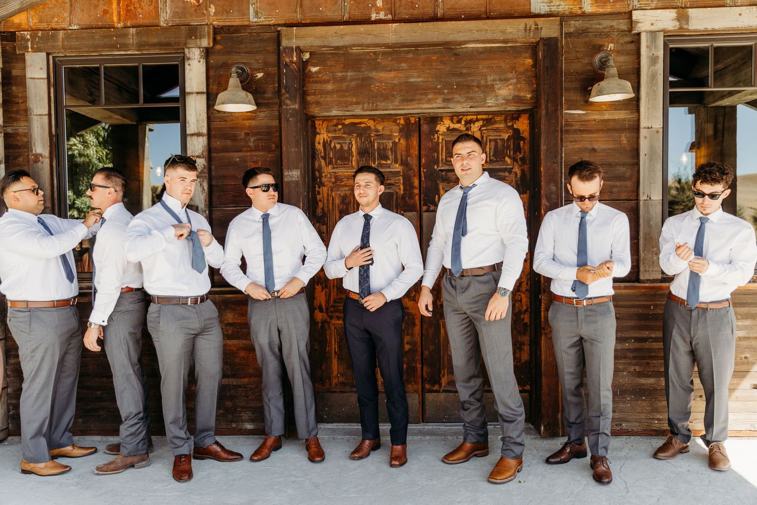 Wedding party groomsmen lined up against rustic wooden wall wearing matching grey suits and blue ties.