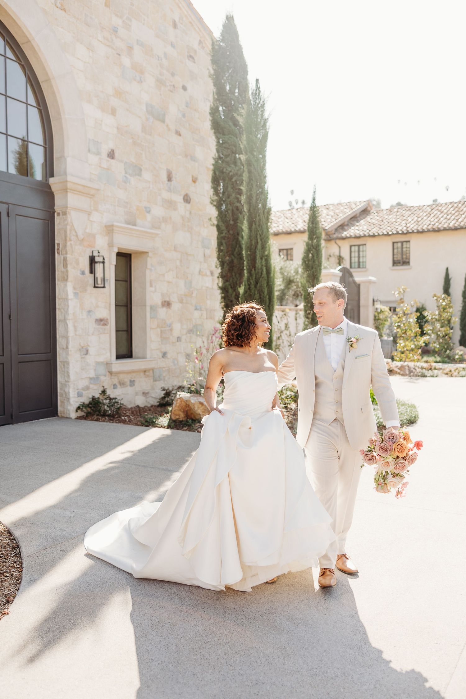 Wedding couple in white attire sitting on stone steps of Mediterranean-style building.