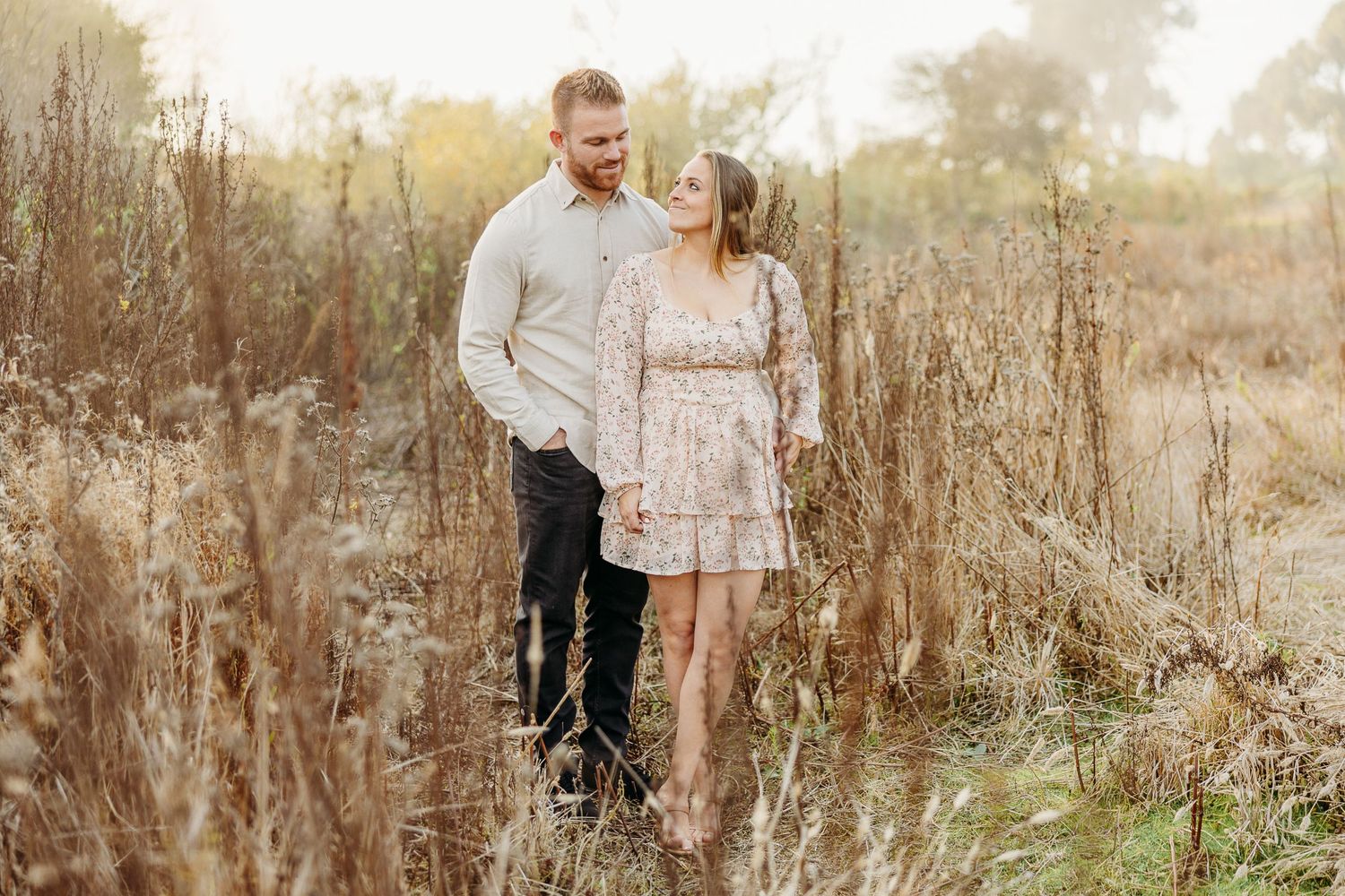 A romantic couple stands together in a golden sunlit field of tall dried grasses at sunset.