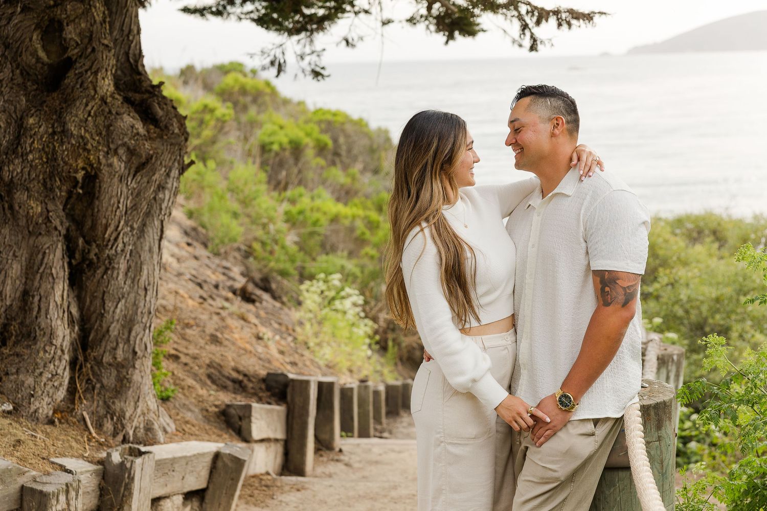 A couple in light colored outfits share a romantic moment on a scenic coastal hiking trail beneath a large tree.