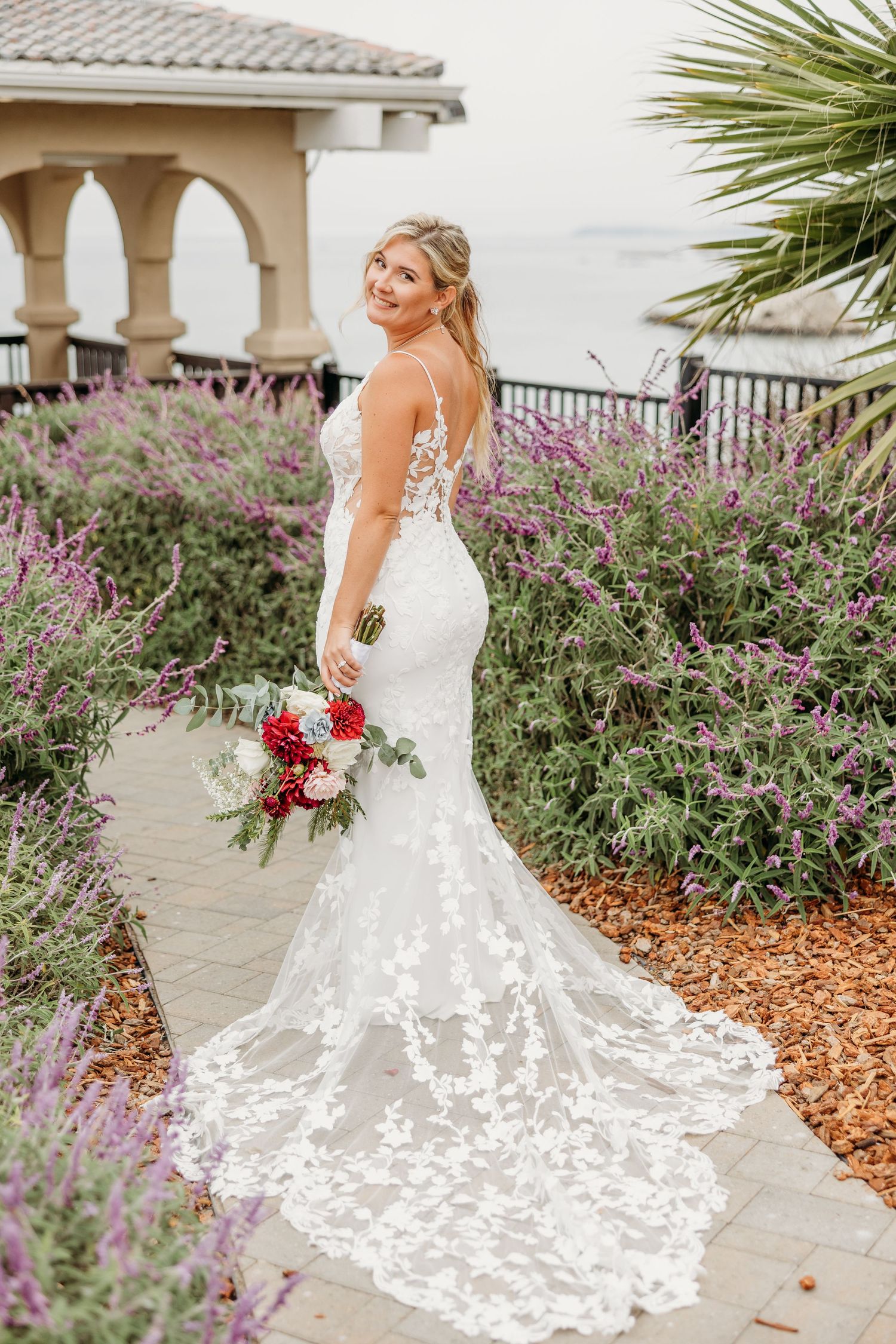 Stunning lace wedding gown with long train photographed among coastal gardens and purple blooms.