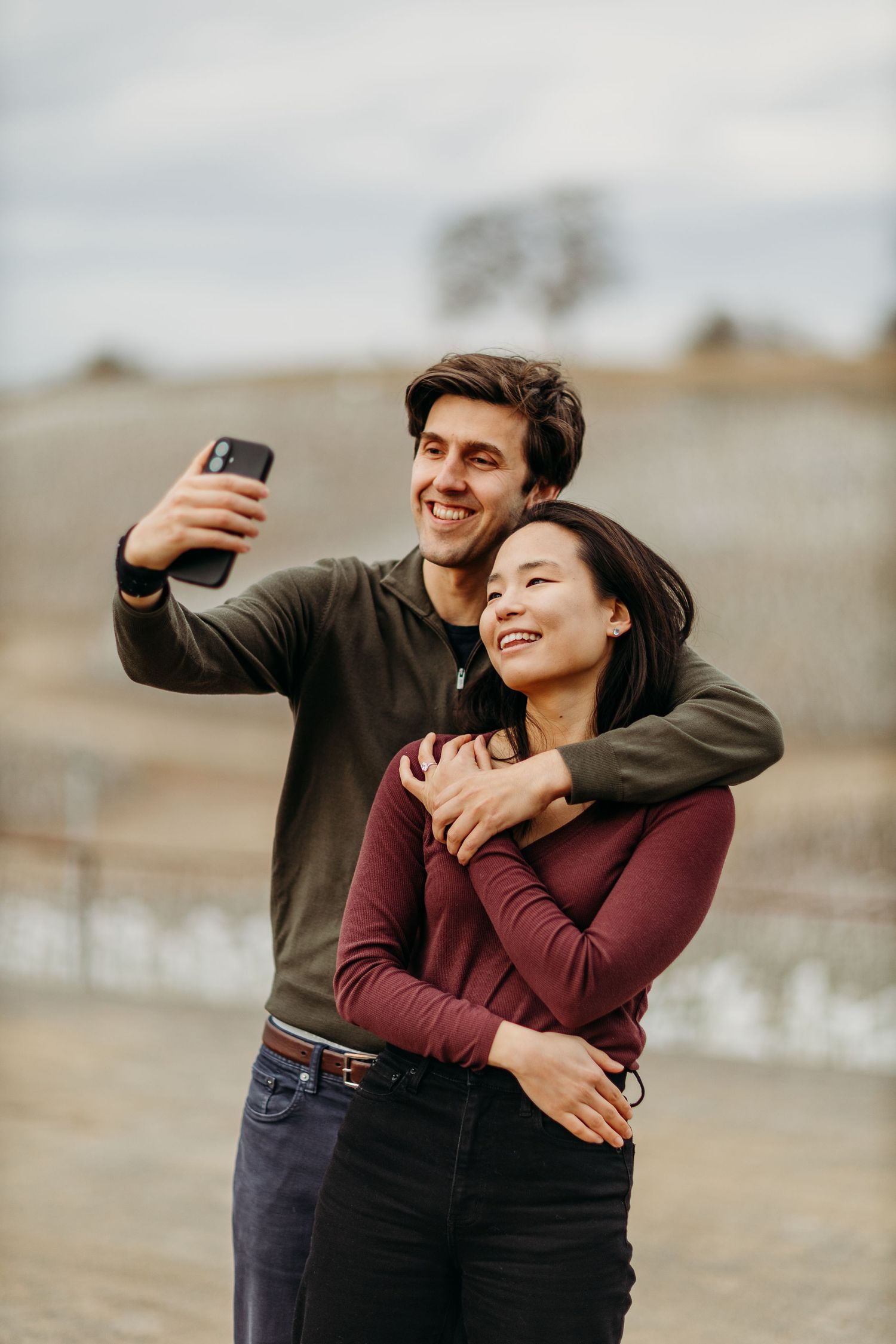 A couple laughs together while taking selfies outdoors on a cloudy day.