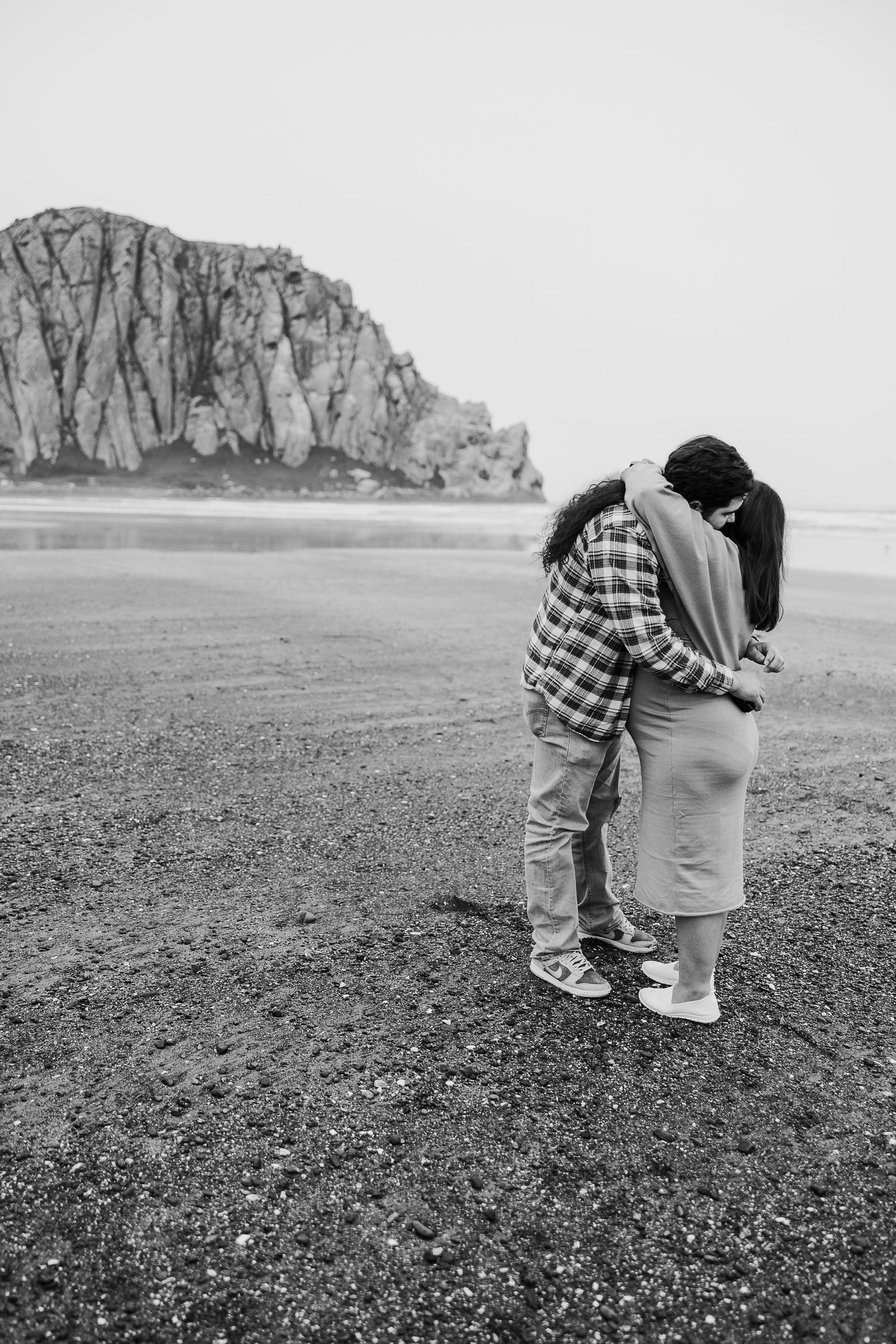 A couple embraces on a rocky beach with Morro Rock in the background, captured in black and white.