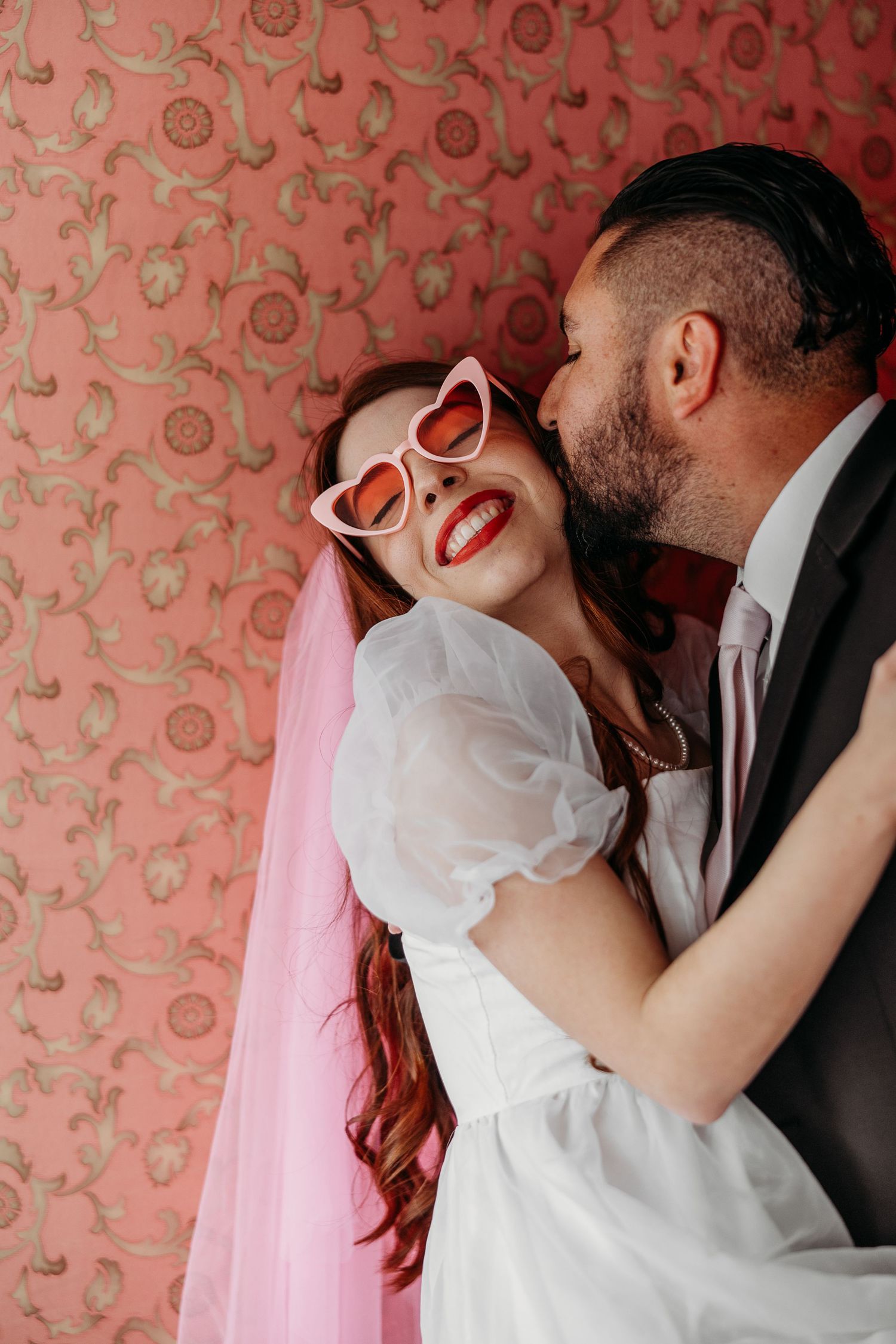A romantic bride in heart-shaped sunglasses and pink hair laughs joyfully against a floral wallpaper backdrop.