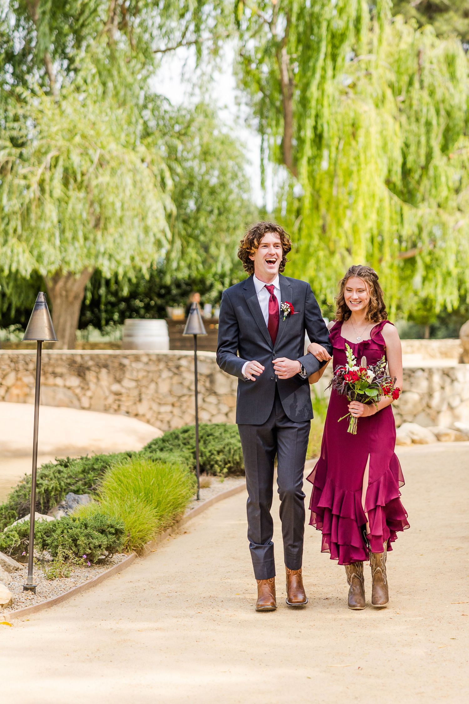 A couple walking down a garden path lined with weeping willow trees.