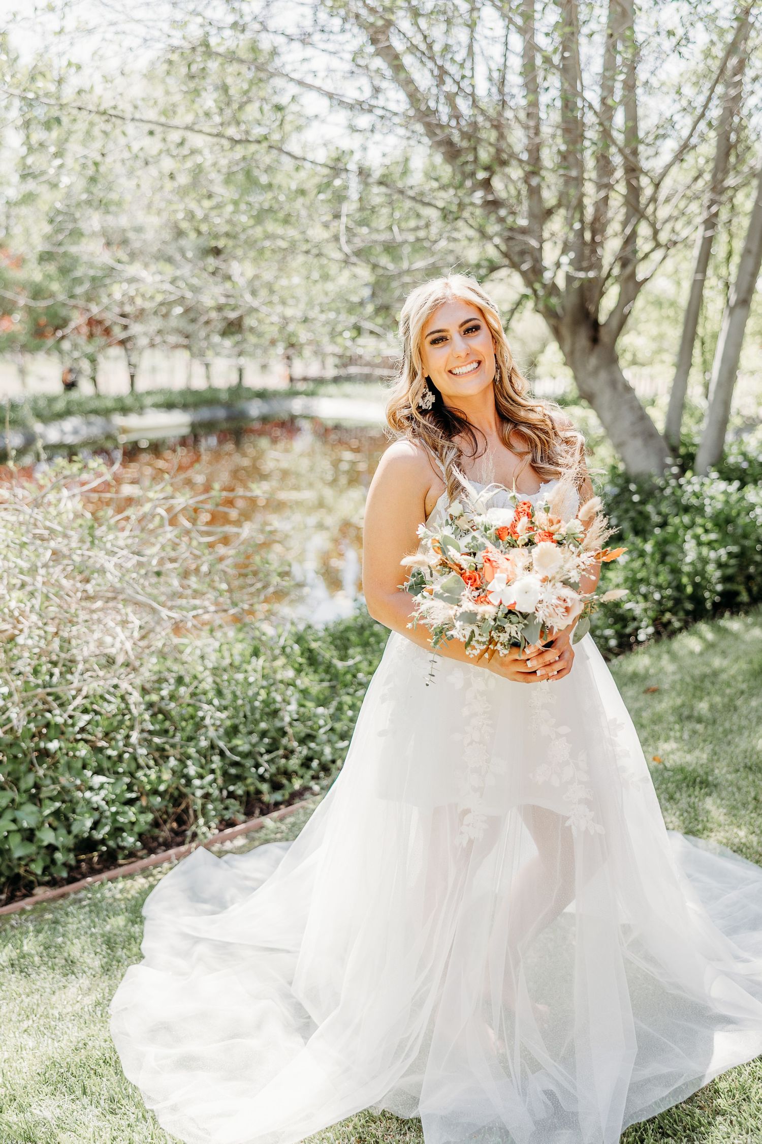 A bride in a flowing white wedding dress holds a peach and white bouquet while standing in a sunny garden setting.