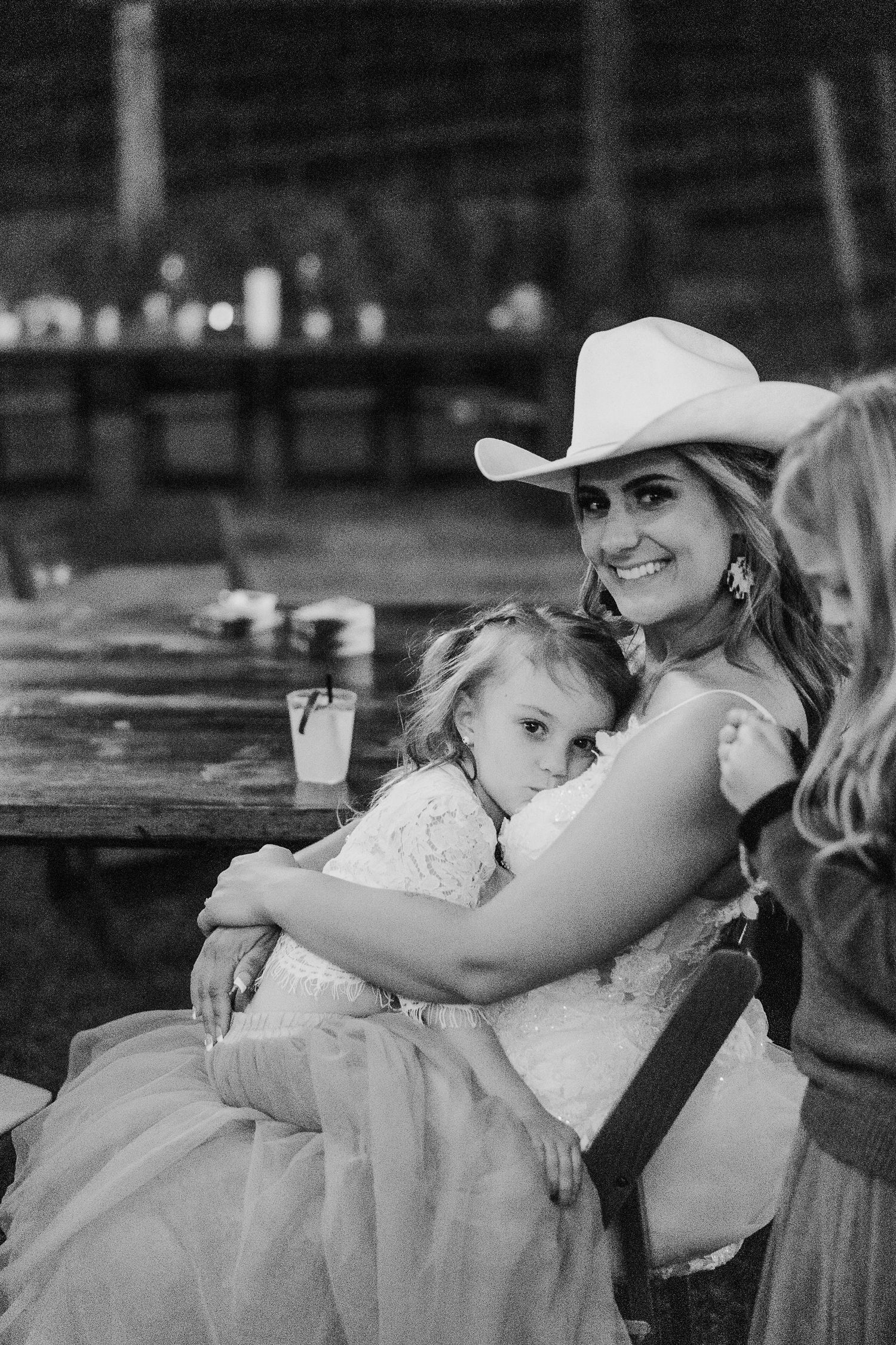 A tender black and white moment shows a mother in a cowboy hat embracing a young child at a rustic wooden table.