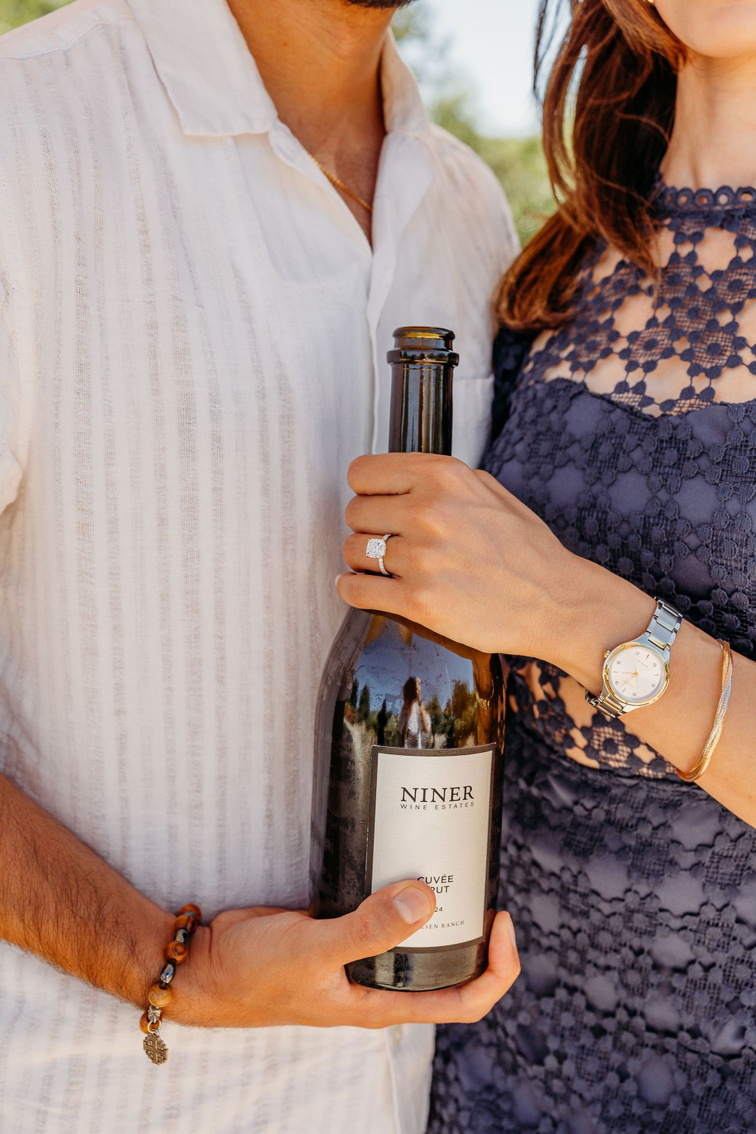Couple holding wine bottle showing engagement ring and watch details with navy lace dress.