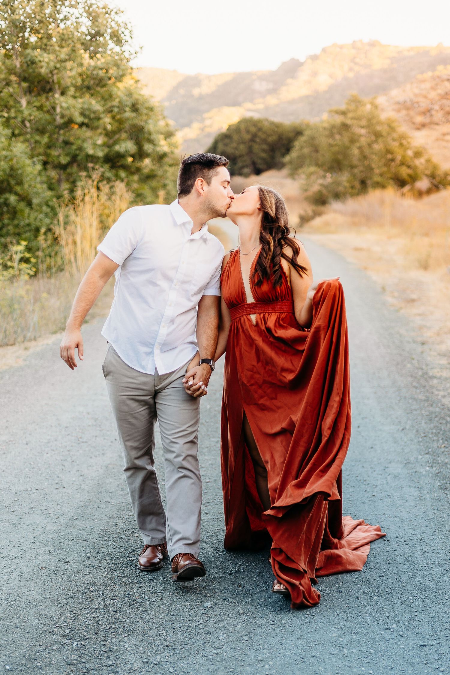 A couple walks hand in hand on a mountain path, with her wearing a flowing red dress and him in casual attire.
