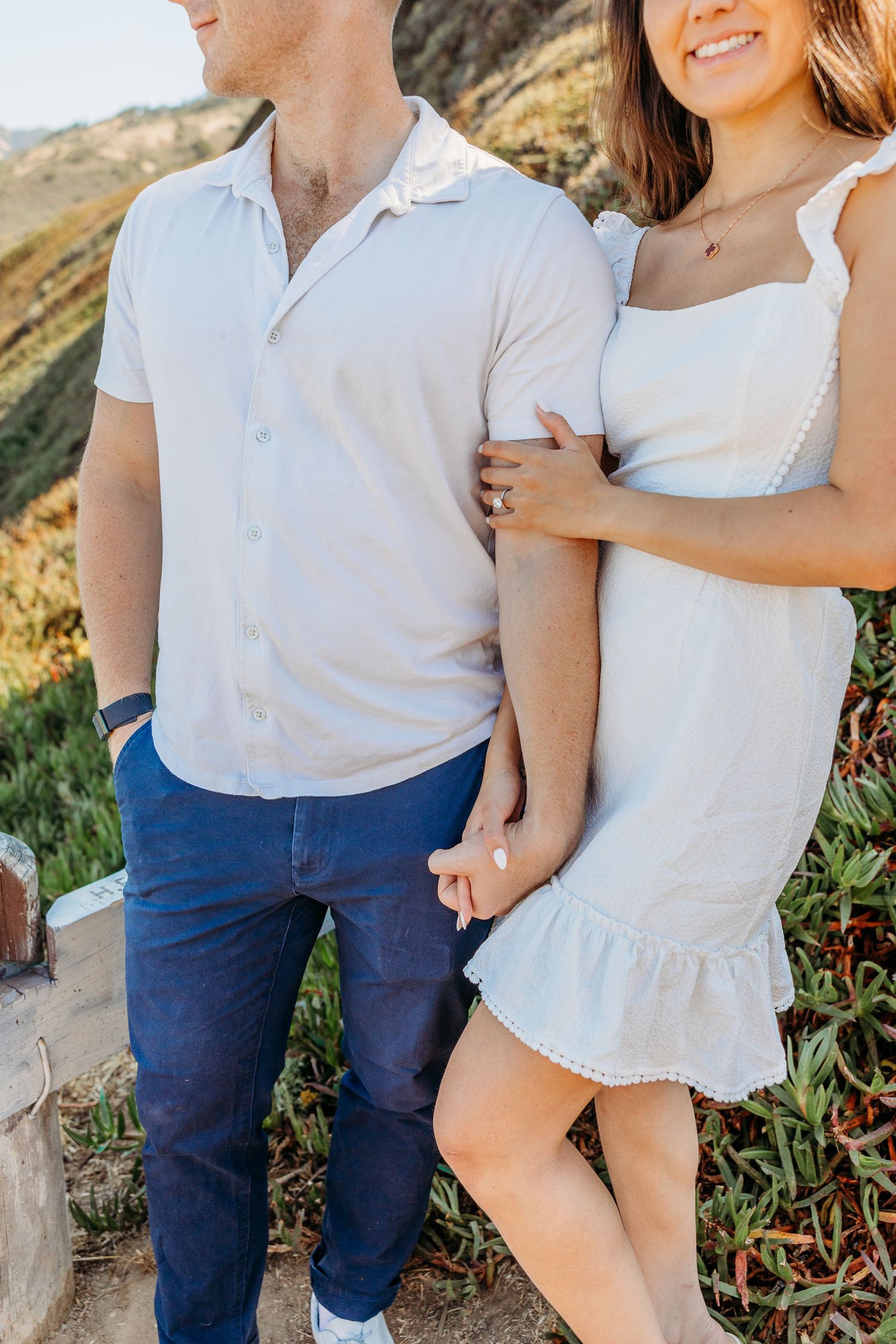 A couple in coordinating white outfits pose together on a coastal cliff.