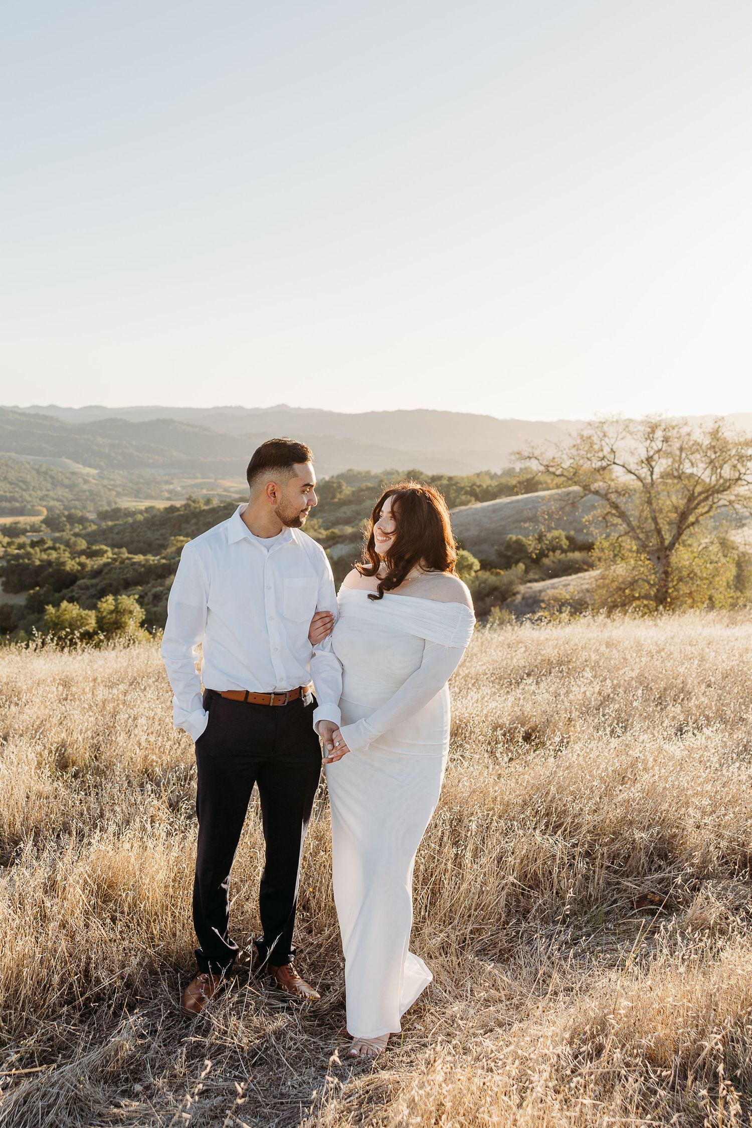 Couple walking together in golden hillside meadow during sunset photoshoot in California landscape.