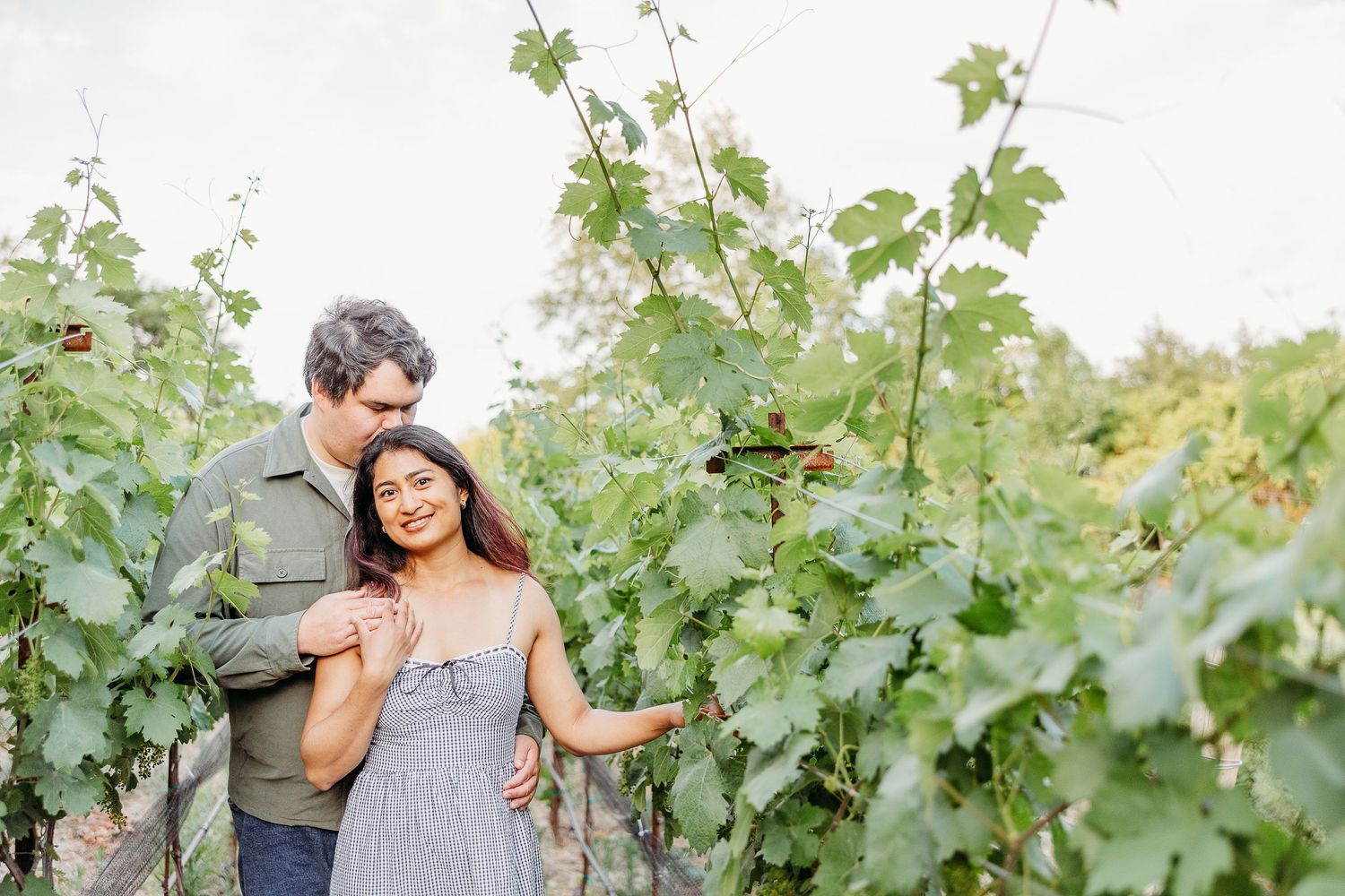 A loving couple smile while walking together through lush green grapevines at a sunny vineyard.