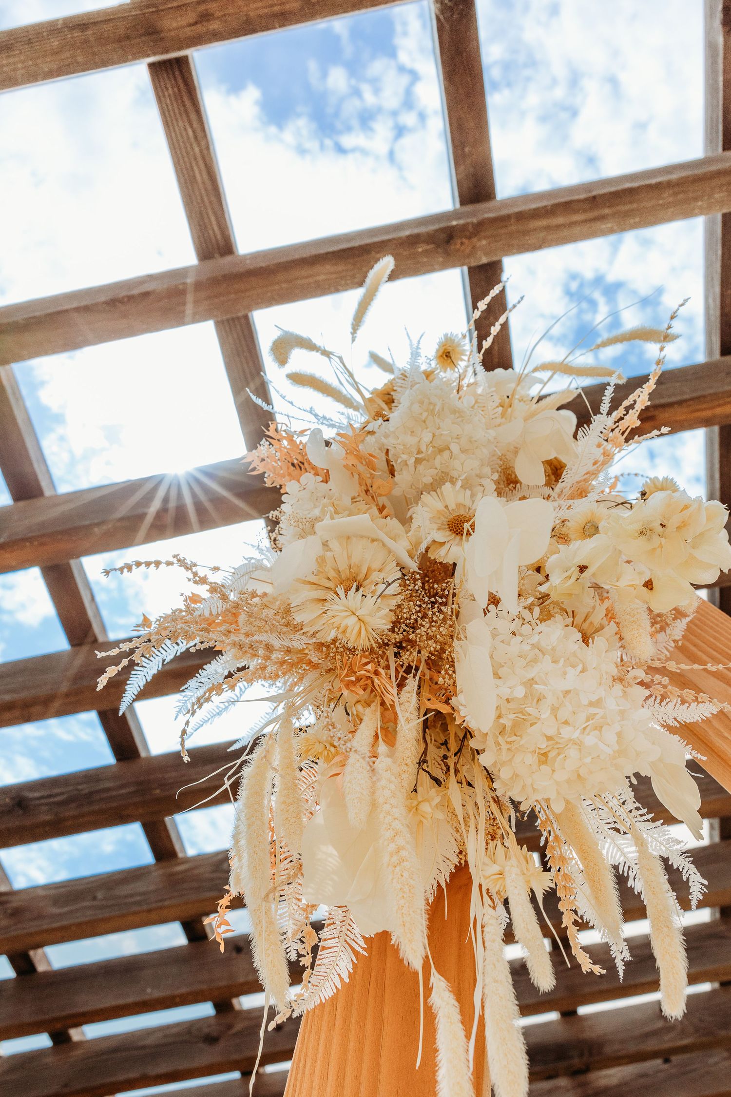 Elegant dried floral arrangement with white blooms under a wooden pergola with blue sky and sunlight peeking through.