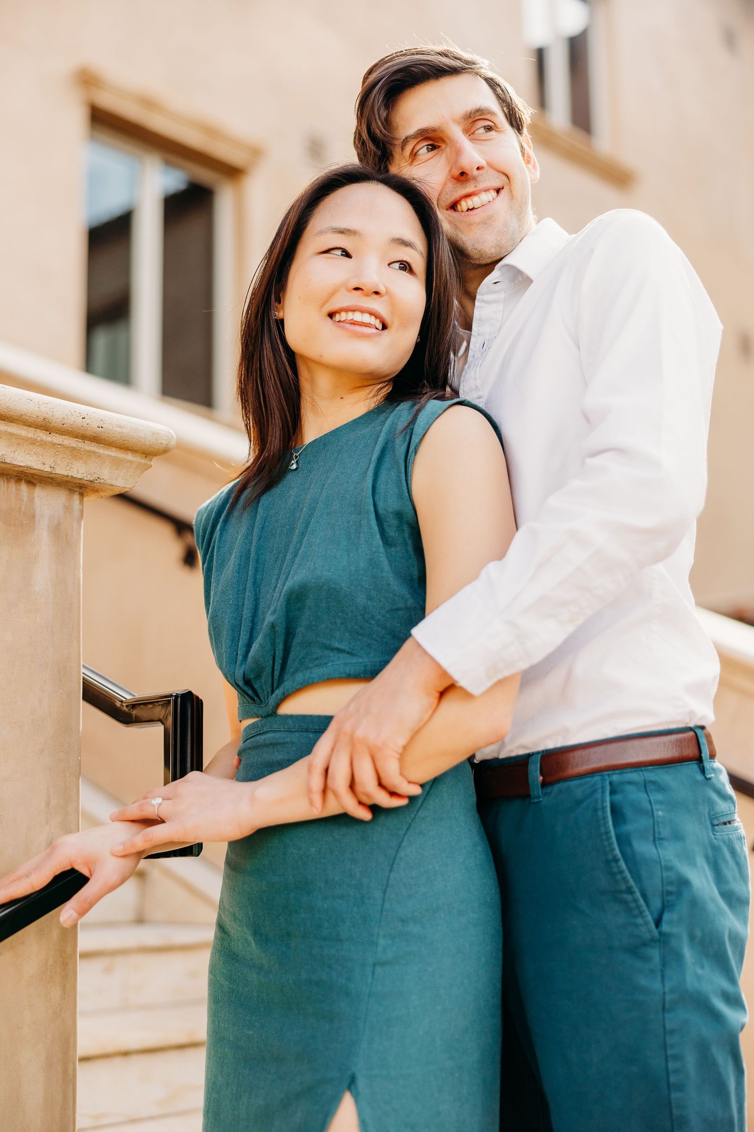 A romantic couple embraces against a beige building exterior while wearing white and teal outfits.