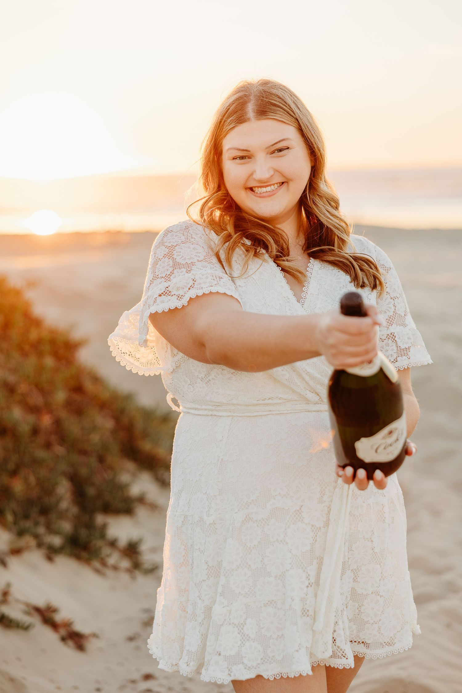 Graduate in white lace dress celebrates with champagne bottle on beach at sunset.