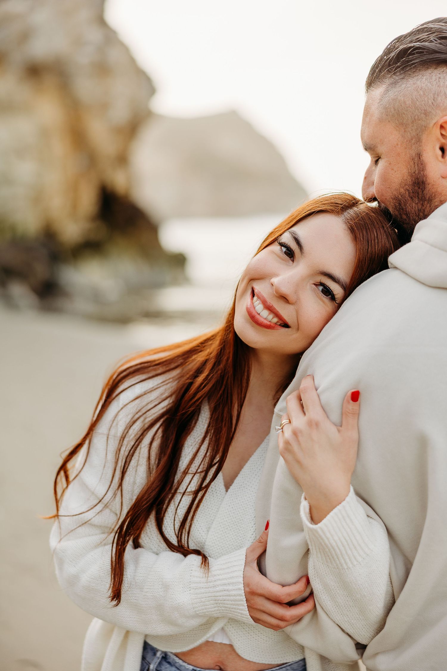 A loving couple embraces tenderly in white sweaters against a soft outdoor background during a romantic photoshoot.