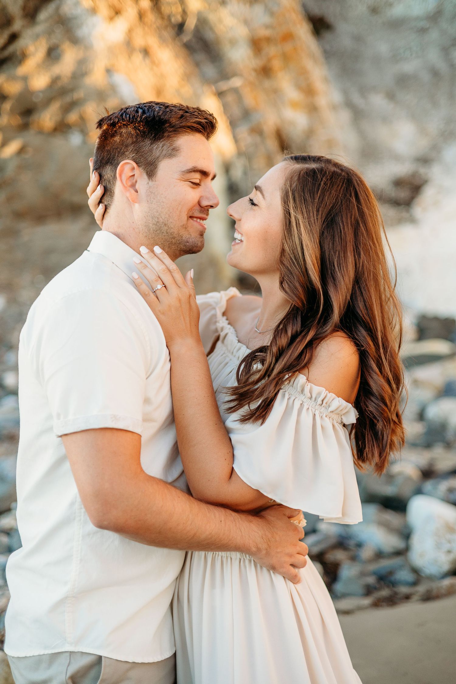 Romantic couple embracing on beach with rocky cliffs in golden sunlight wearing white summer outfits.