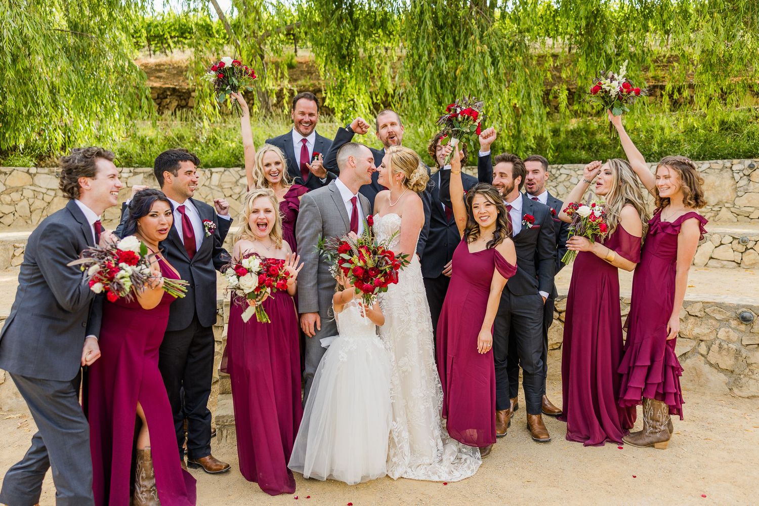 Celebratory wedding party photo with burgundy and gray attire against stone wall backdrop.