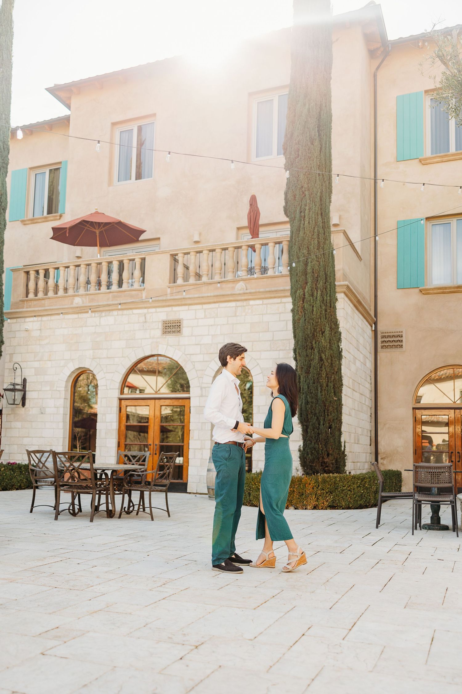 A couple stands together outside a Mediterranean-style building with cypress trees and sunlit architecture.