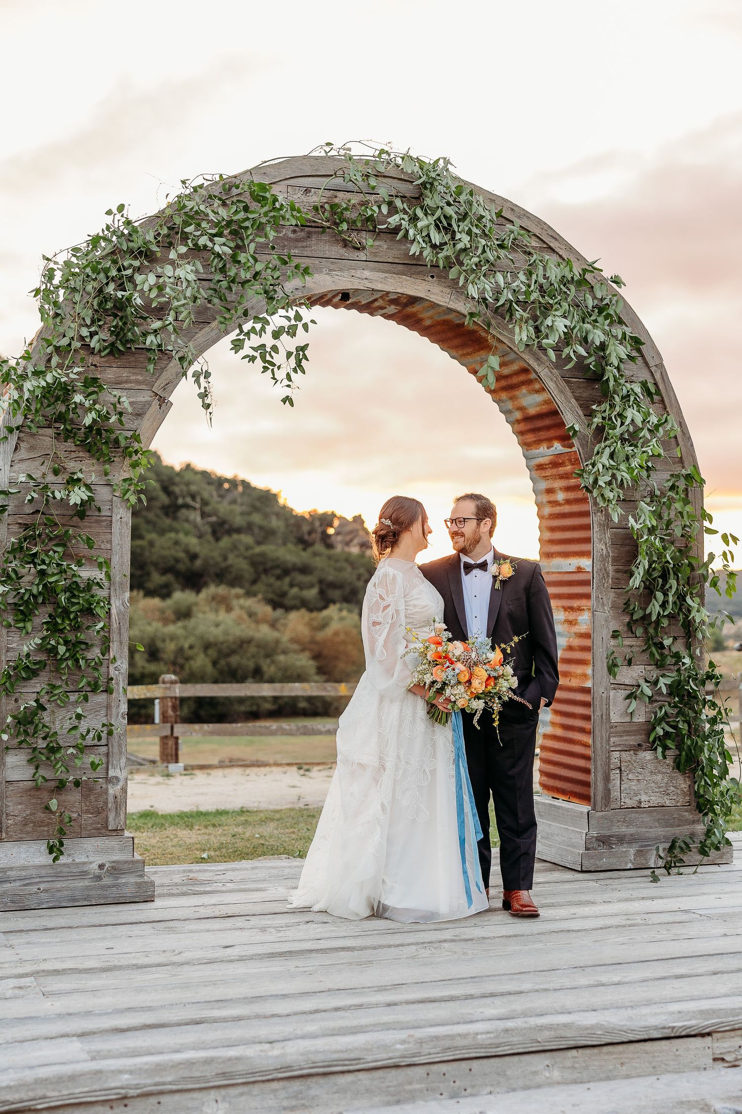 Romantic wedding couple stands under an ivy-covered brick archway at sunset holding a vibrant autumn bouquet.
