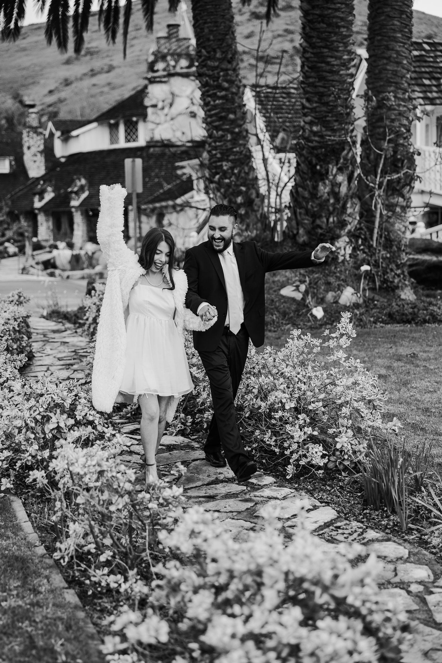 A couple walks down a garden path lined with flowers and palm trees in black and white photography.