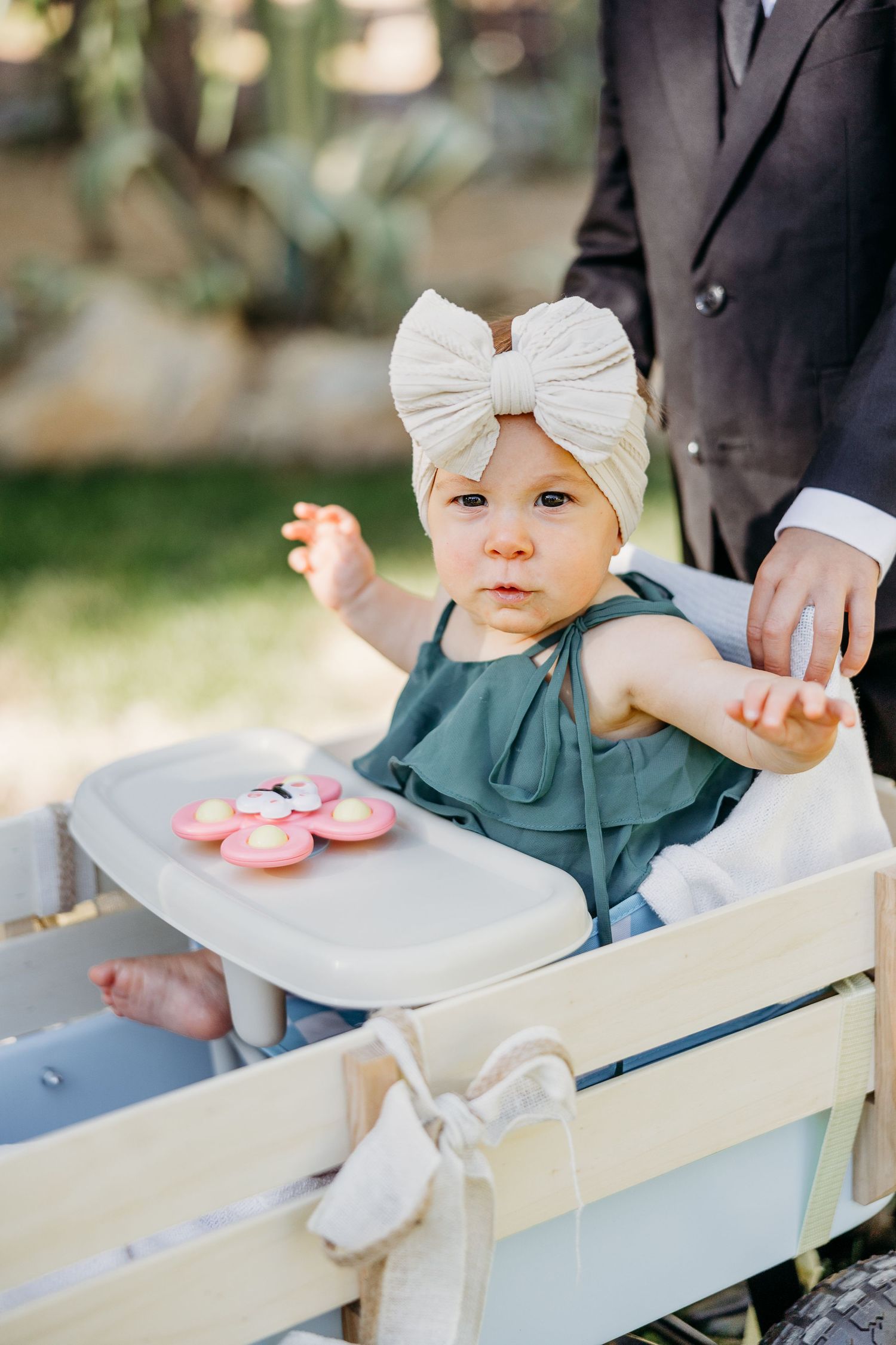 Baby wearing white bow headband and blue dress sits in wooden wagon with pink shoes on tray.