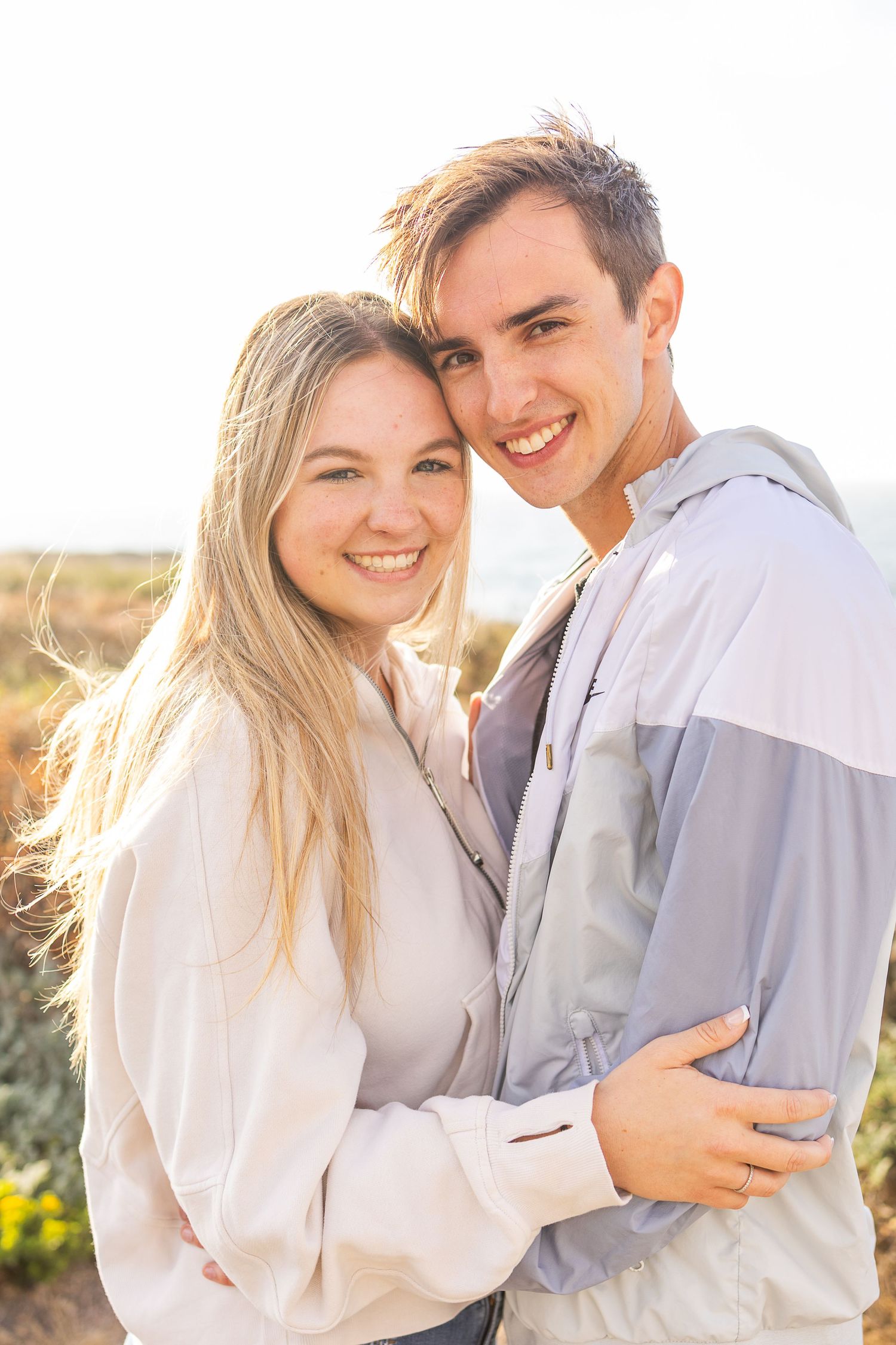 A couple in matching light hoodies poses together against an ocean backdrop.