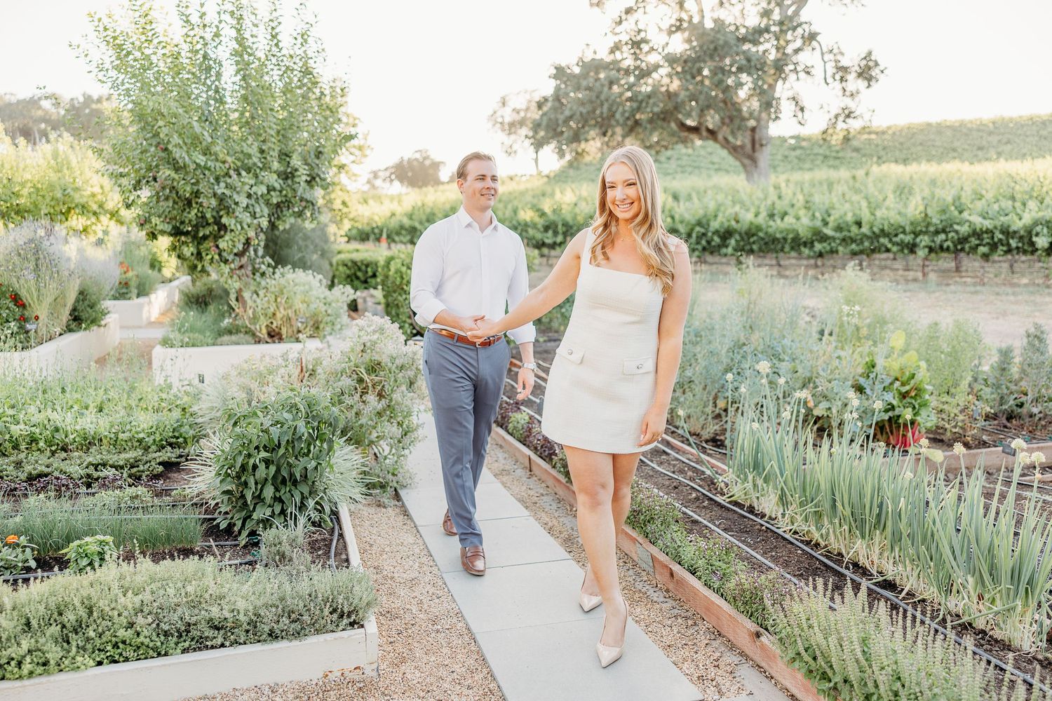 A couple walks together through a lush garden path with blooming flowers and greenery at sunset.